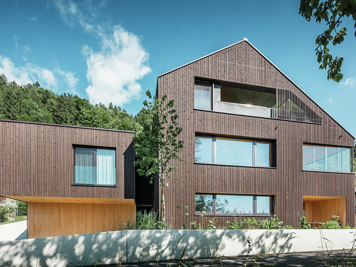 Un immeuble moderne avec un toit PREFA Prefalz brun noisette et une façade en bois sombre, parsemée d'éléments en bois clair, de grandes fenêtres et entourée d'un paysage verdoyant.
