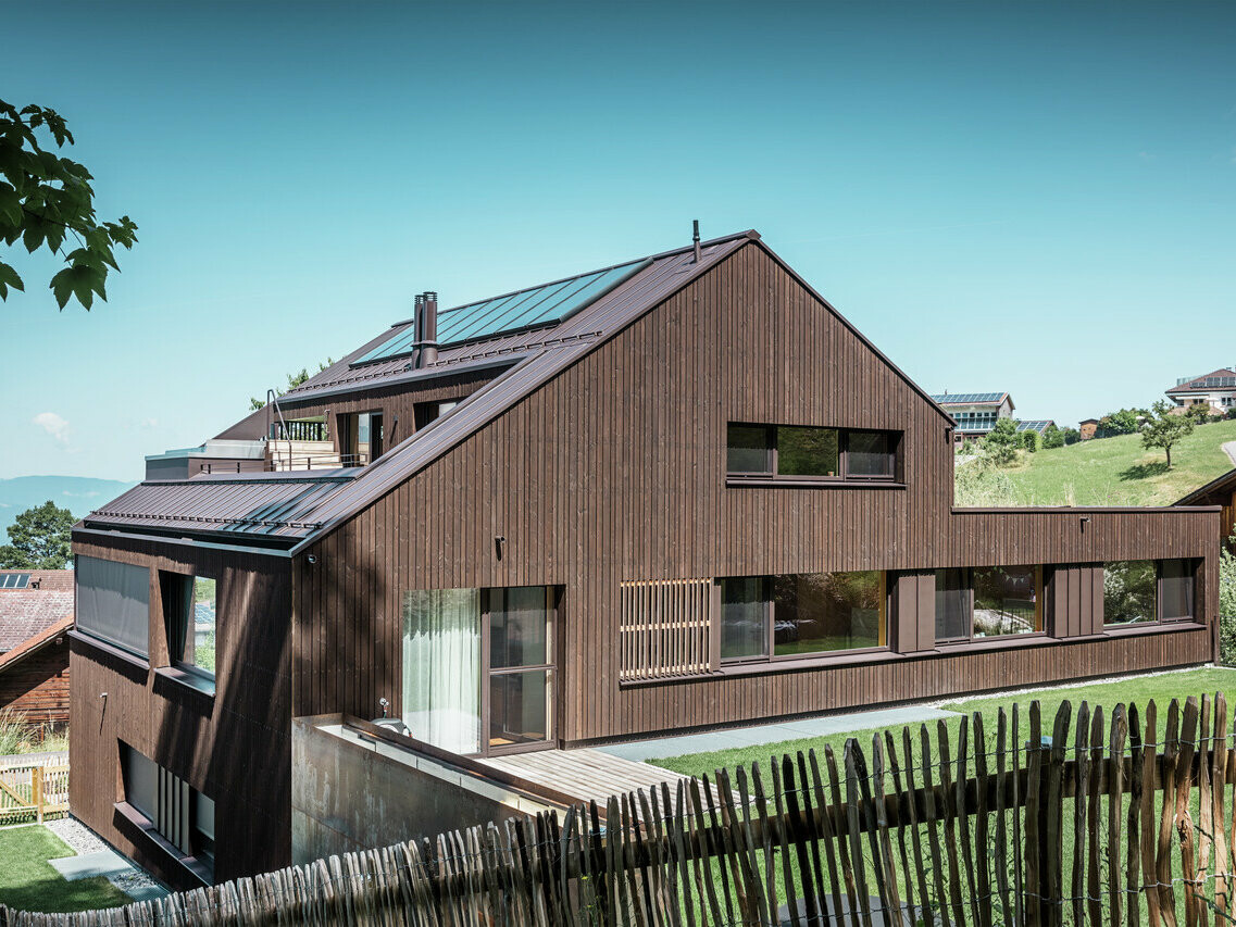 Un immeuble moderne dans le Vorarlberg, en Autriche, avec un toit à deux versants recouvert de Prefalz en brun noyer. La façade en bois du bâtiment s'intègre harmonieusement dans l'environnement rural, tandis que de grandes fenêtres apportent une touche de modernité.