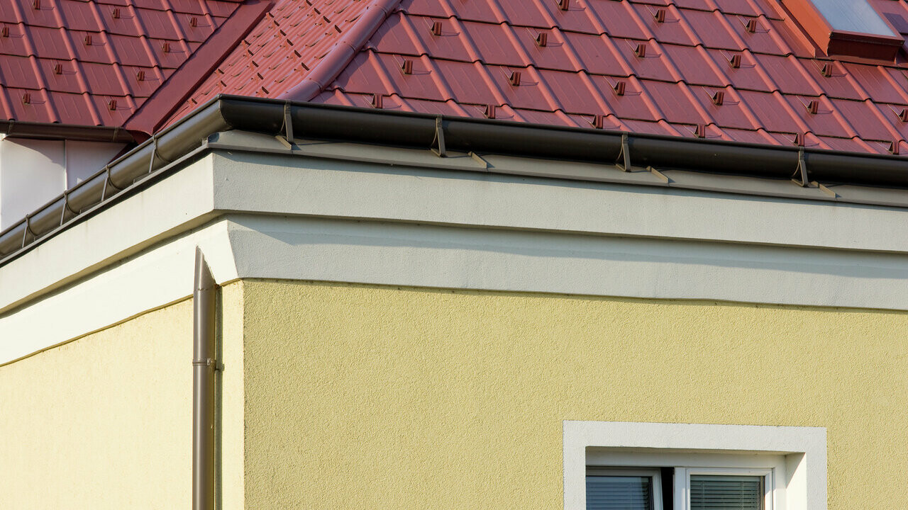Hoek van een huis met gele pleistergevel en bruine PREFA Ardense goot (op het dak gemonteerde goot) met rode PREFA dakpannen.