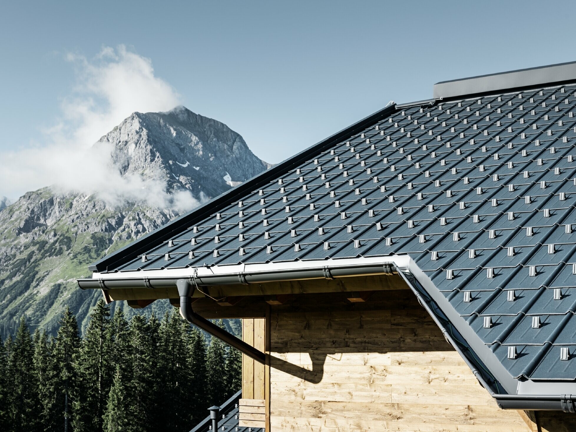 PREFA-dakpan in een bergstreek - dakplaat in antraciet, gemonteerd op de berghut in Lech am Arlberg met bergen op de achtergrond; PREFA-dakgoot in antraciet; houten gevel