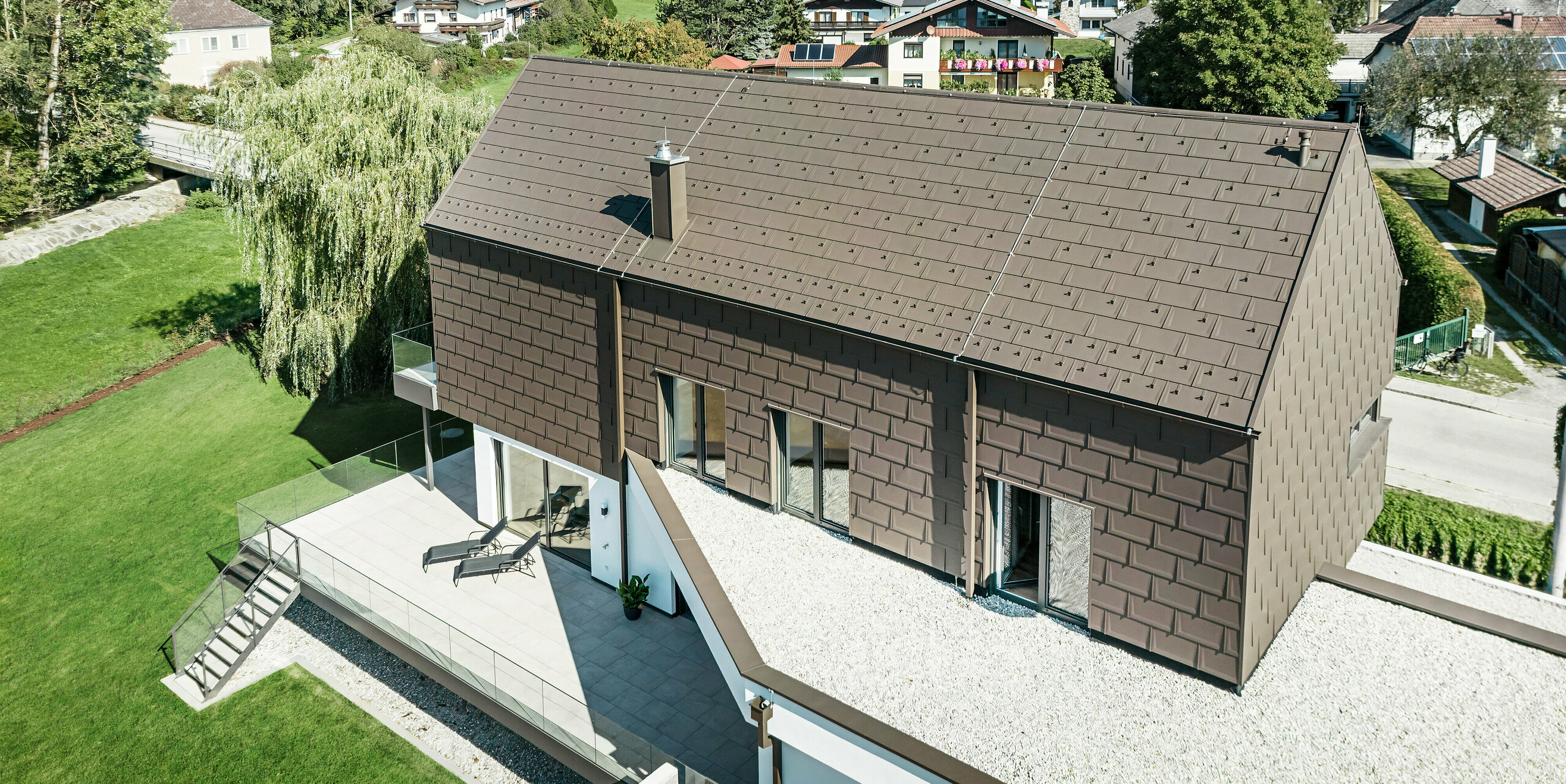 Blick aus der Vogelperspektive auf ein einzigartiges Einfamilienhaus in Neukirchen, Österreich, eingedeckt mit PREFA R. 16 Dachplatten in P.10 braun. Kleinformatige Aluminiumelemente zieren das Satteldach und das Obergeschoss des modernen Hauses. Die Dachentwässerung aus PREFA in der gleichen Farbe erfolgt über ein Vierkantrohr und eine Kastenrinne mit Wassersammelkasten. Die großen Fenster geben den Blick aus dem Inneren des Gebäudes auf die ruhige Wohnlandschaft von Neukirchen mit ihren zahlreichen grünen Baumkronen frei.