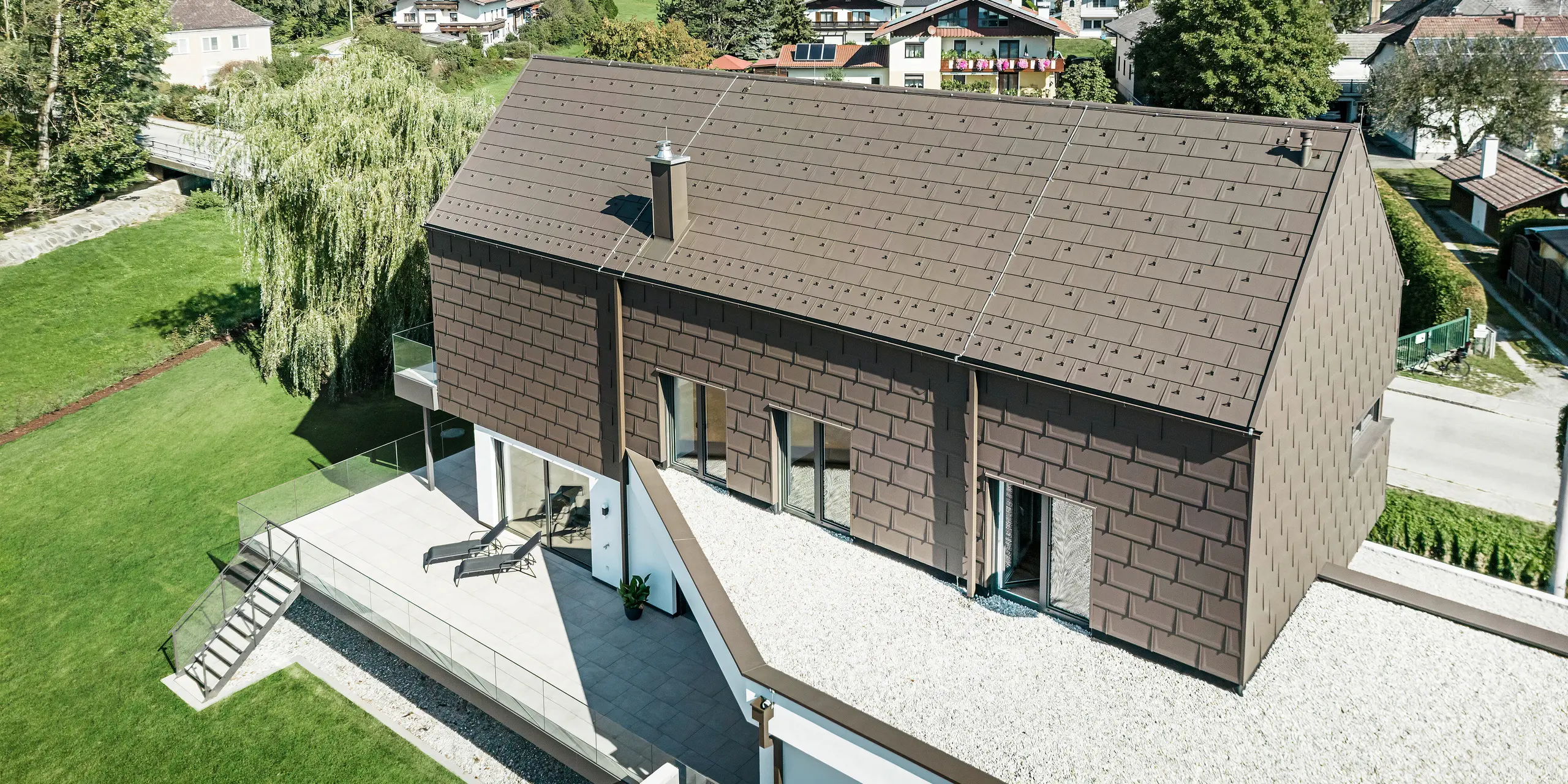 Blick aus der Vogelperspektive auf ein einzigartiges Einfamilienhaus in Neukirchen, Österreich, eingedeckt mit PREFA R. 16 Dachplatten in P.10 braun. Kleinformatige Aluminiumelemente zieren das Satteldach und das Obergeschoss des modernen Hauses. Die Dachentwässerung aus PREFA in der gleichen Farbe erfolgt über ein Vierkantrohr und eine Kastenrinne mit Wassersammelkasten. Die großen Fenster geben den Blick aus dem Inneren des Gebäudes auf die ruhige Wohnlandschaft von Neukirchen mit ihren zahlreichen grünen Baumkronen frei.