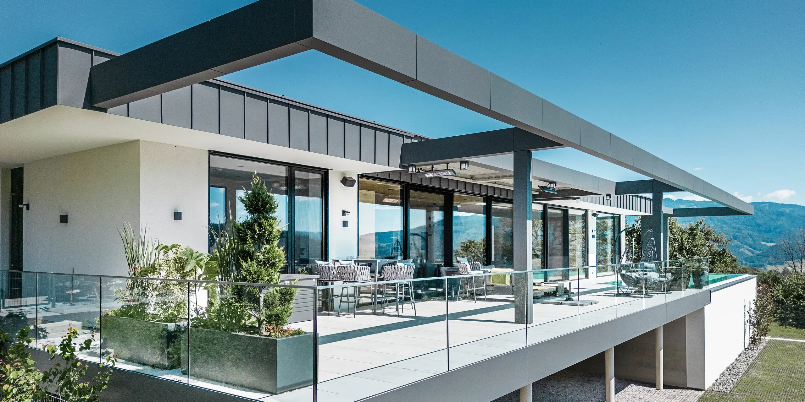 Luxuriöse Terrasse eines Einfamilienhauses in Pogier, Österreich mit eleganten Dach- und Fassadensystemen von PREFA. Die Kombination von PREFALZ und PREFABOND in dunkelgrau ergibt ein modernes und anspruchsvolles Design. Die großzügige Terrasse bietet einen atemberaubenden Ausblick und ist mit einem Glasgeländer ausgestattet, das eine offene und einladende Atmosphäre schafft. Der teilweise überdachte Außenbereich lässt die Schönheit der umliegenden Natur genießen und ist ideal zum Entspannen und geselligen Beisammensein.