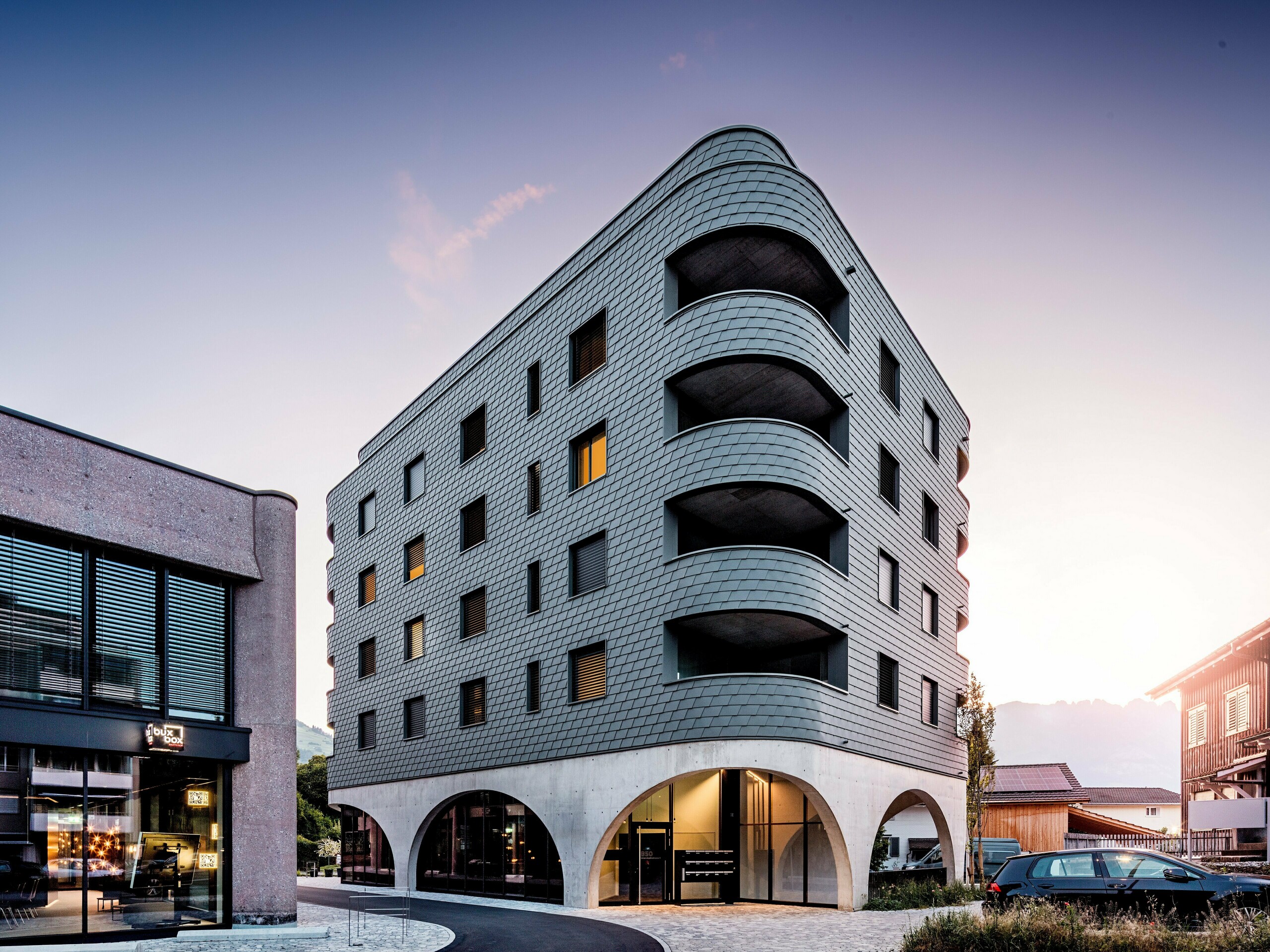 Modernes Mixed-Use-Gebäude in Buchs mit markant gerundeter, dreieckiger Form und durchgehender Fassade aus PREFA Wandschindeln in P.10 Hellgrau. Die schuppenartige Aluminiumhaut umhüllt das Gebäude wie eine Reptilienstruktur und betont die fließenden Rundungen sowie die tief eingeschnittenen Balkone. Über dem massiven Sockel mit eleganten Betonarkaden wirkt die leichte, witterungsbeständige PREFA Fassade besonders ausdrucksstark. Präzise verlegte Aluminiumschindeln sorgen für eine homogene, langlebige und wartungsarme Gebäudehülle mit starkem Wiedererkennungswert.