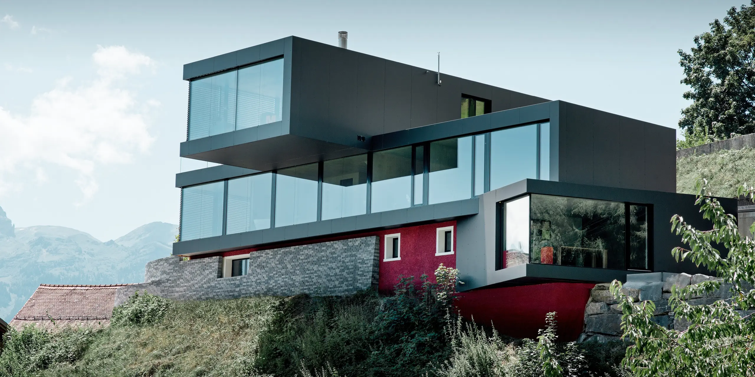 Spektakuläres Einfamilienhaus in Walenstadtberg mit markanter Fassade aus schwarzgrauen PREFA Aluminium Verbundplatten vor beeindruckender Bergkulisse. Die kubische Architektur mit weit auskragenden Baukörpern und großflächigen Glasfronten wirkt wie ein modernes Designobjekt in der Landschaft. Die langlebige, wartungsarme Aluminiumfassade begeistert durch klare Linien, hohe Witterungsbeständigkeit und zeitlose Eleganz.