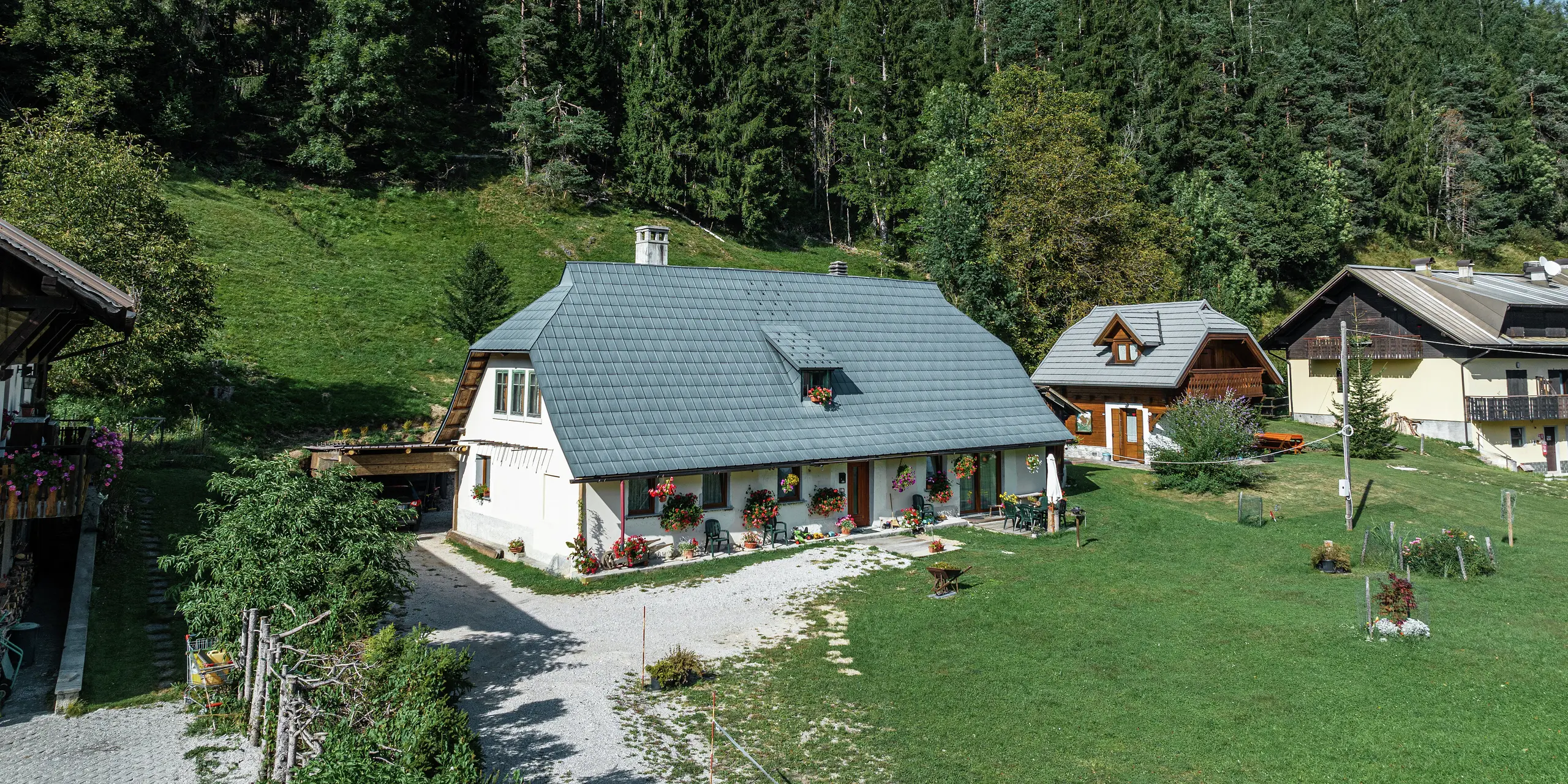 Vue aérienne d'une maison familiale idyllique à Tarvisio, en Italie, protégée par des tuiles PREFA durables depuis 1972. Le grand toit en aluminium résistant aux intempéries s'intègre harmonieusement dans le paysage alpin verdoyant. La façade joliment décorée de fleurs et la terrasse chaleureuse confèrent à la maison une atmosphère accueillante. Entourée de maisons traditionnelles aux façades en bois et d'une forêt dense de conifères, la propriété offre un mélange parfait de proximité avec la nature et de stabilité architecturale.