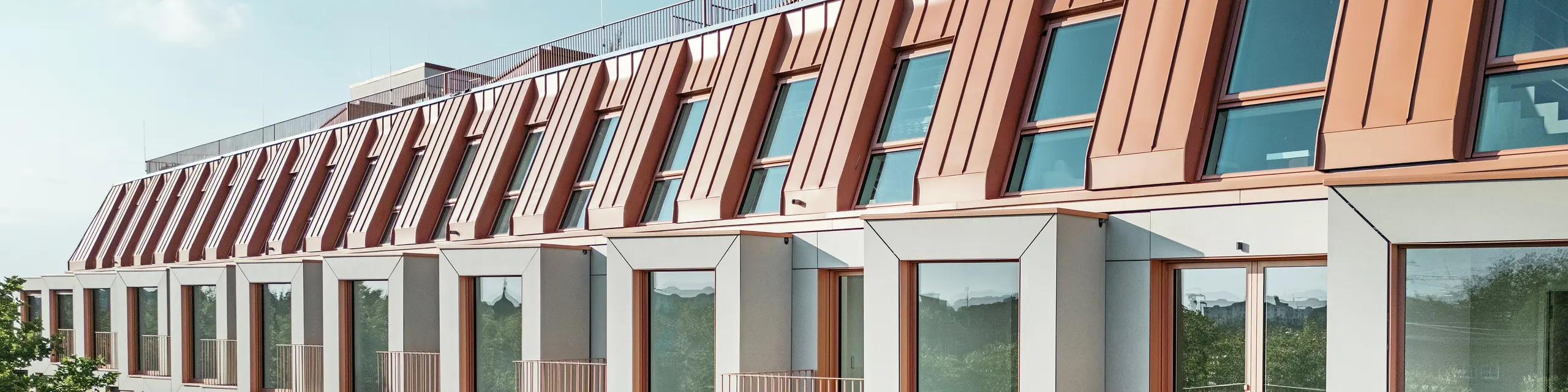 Vue de la façade du projet résidentiel Van B à Munich avec le toit en aluminium PREFALZ remarquable dans la couleur spéciale IGP Orange HDP de PREFA. La couverture de toit moderne allie élégance esthétique et précision technique et offre une protection durable contre les éléments. Les baies vitrées de conception cubique avec de généreuses surfaces vitrées et des balcons délicats créent une interaction harmonieuse entre lumière, espace et fonctionnalité - idéale pour des concepts de vie sophistiqués dans un environnement urbain.