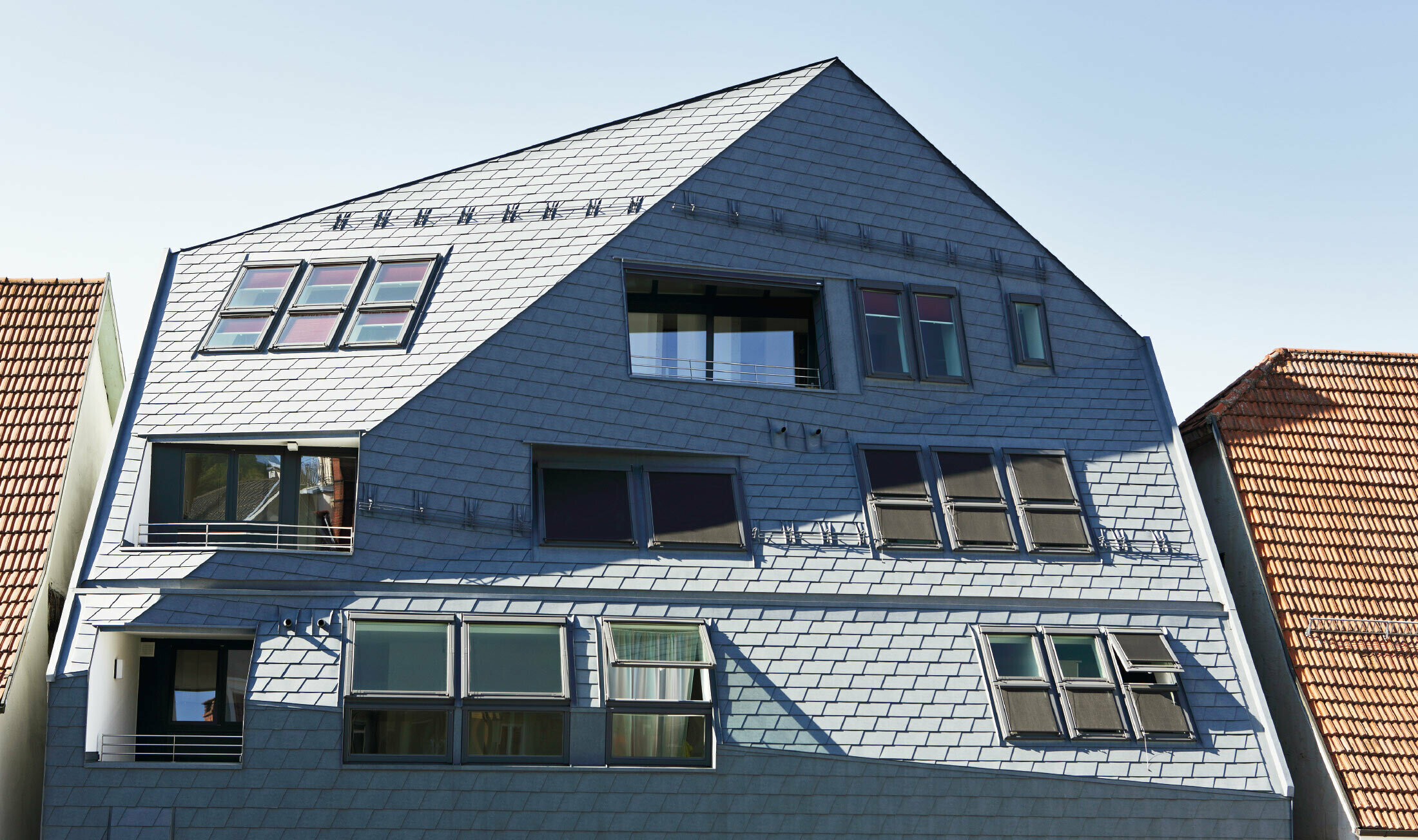 Maison galerie avec façade plissée, revêtue du bardeau de toiture et de façade PREFA en P.10 gris pierre avec de nombreuses fenêtres