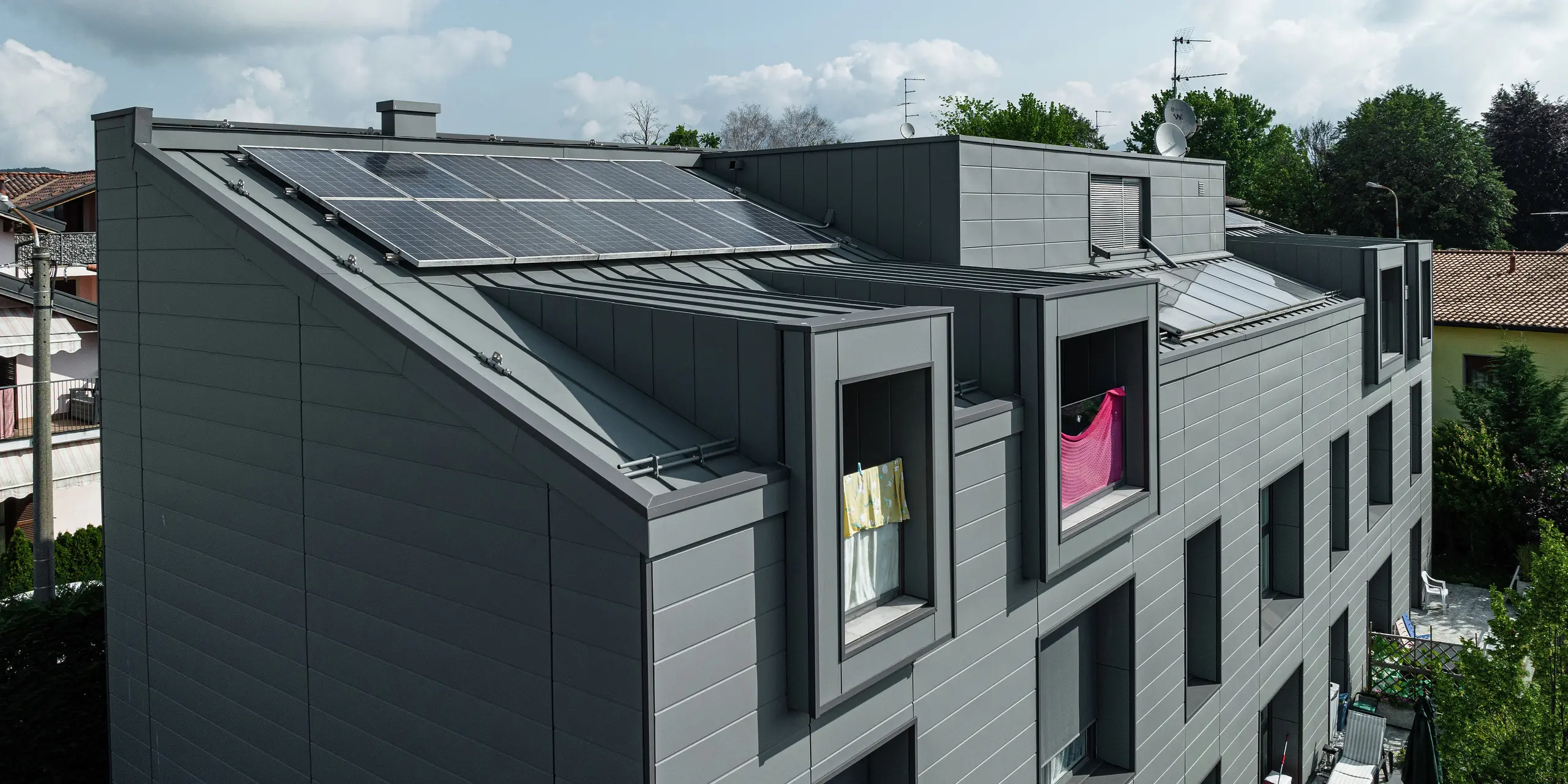 Vue aérienne du toit d'un complexe résidentiel moderne à Faloppio, en Italie, avec un revêtement robuste en aluminium à joint debout et un système photovoltaïque durable. La façade en bardage PREFA et le toit métallique offrent non seulement des points forts visuels, mais également une excellente résistance aux intempéries et une source d'énergie durable grâce aux modules photovoltaïques. La combinaison de produits PREFA fonctionnels et esthétiques garantit une solution durable et économe en énergie.