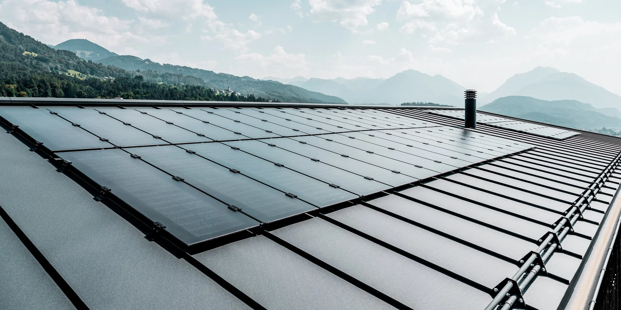 Diese beeindruckende Nahaufnahme zeigt die großflächigen Solarmodule PREFALZ auf einem Einfamilienhaus in Röthis in Vorarlberg. Die Solarmodule fügen sich harmonisch in das PREFALZ Aluminiumdach in der Farbe P.10 Anthrazit ein. Die klaren Stehfalze unterstreichen die moderne Optik, während die leichte, langlebige und witterungsbeständige Dachdeckung zuverlässigen Schutz bietet. Im Hintergrund erstreckt sich die malerische Vorarlberger Berglandschaft.