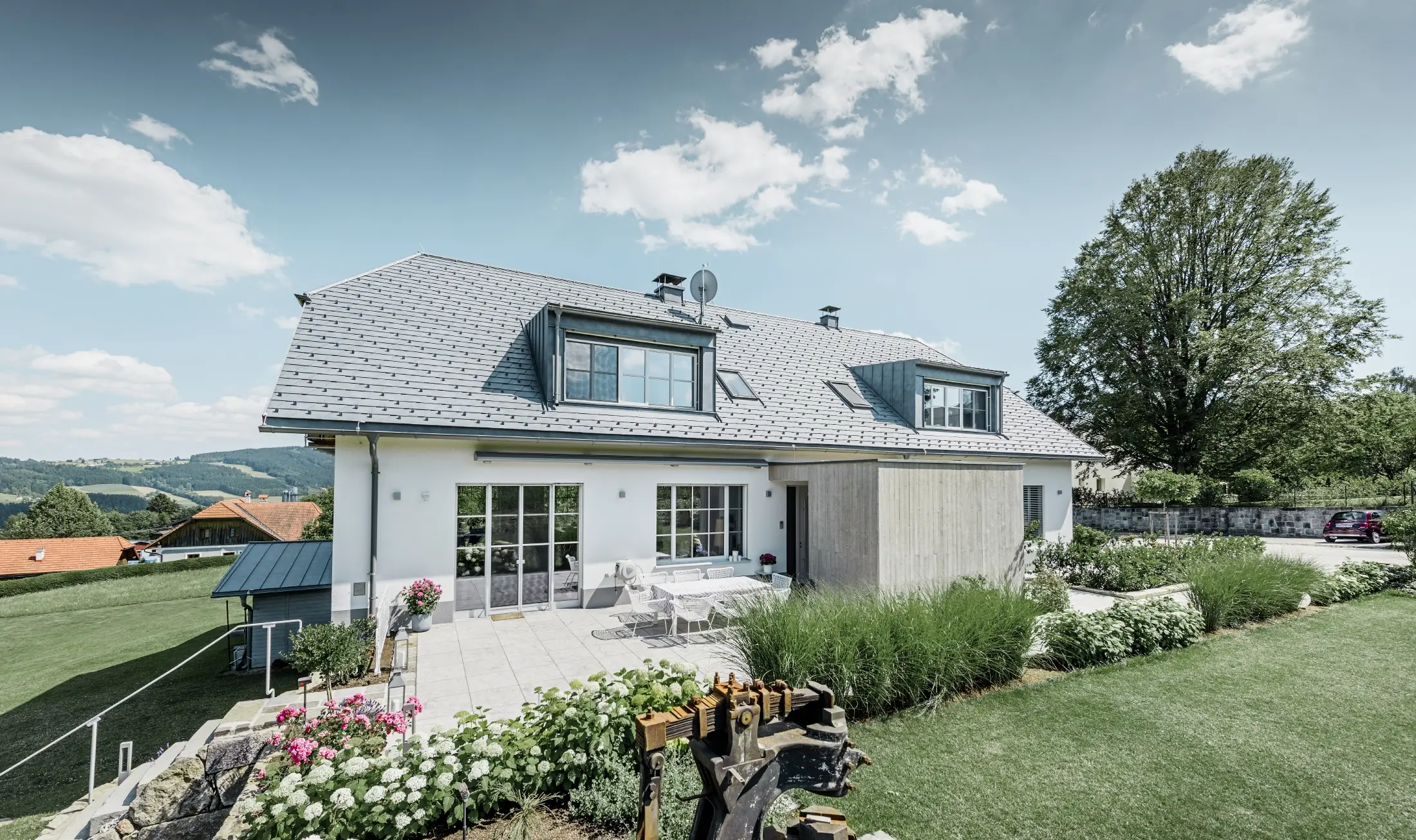 Maison individuelle classique avec toit en demi-croupe ; maison avec toit rénové à l'aide de bardeaux PREFA couleur gris pierre, avec beau jardin et grande terrasse.