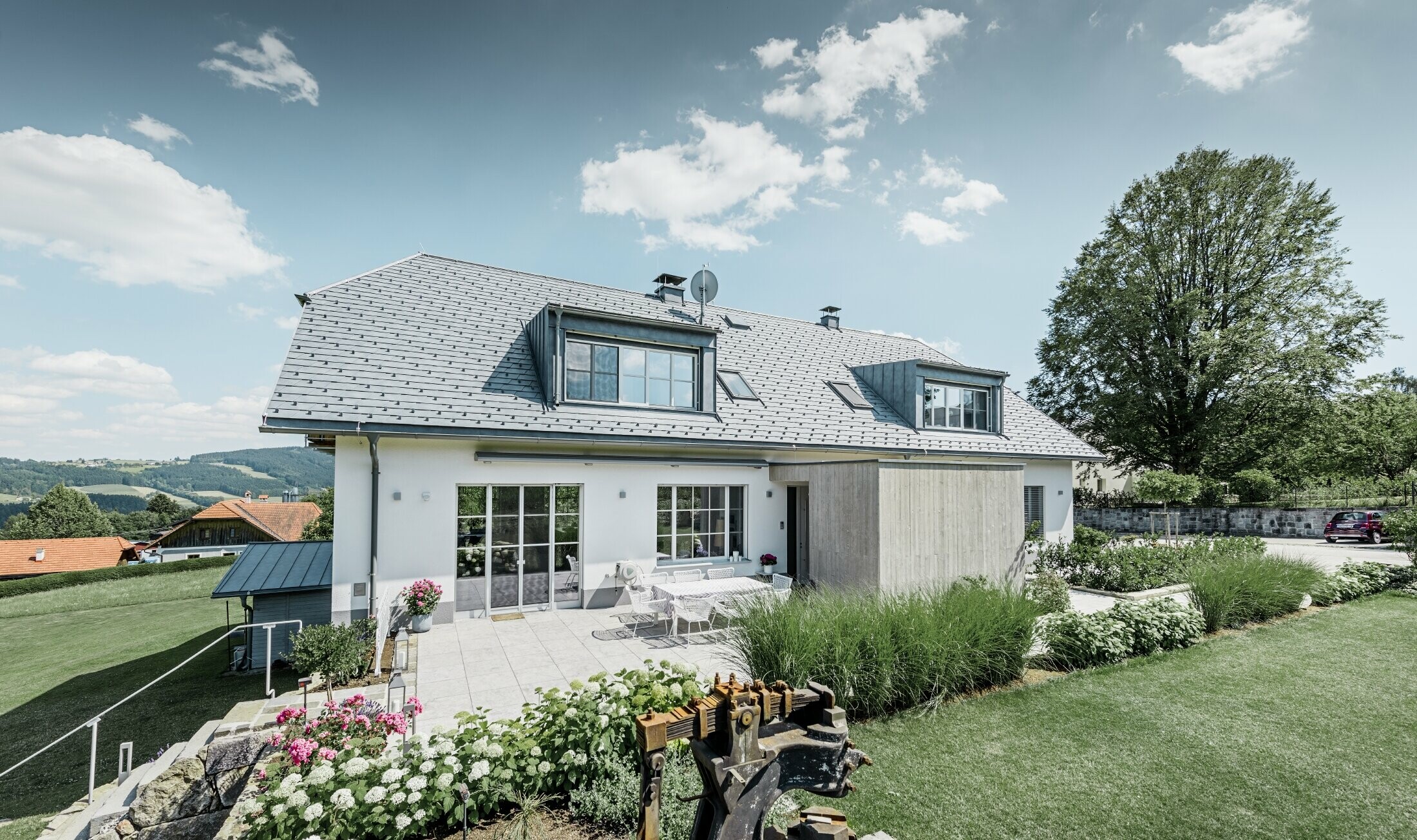 Maison individuelle classique avec toit en demi-croupe ; maison avec toit rénové à l'aide de bardeaux PREFA couleur gris pierre, avec beau jardin et grande terrasse.