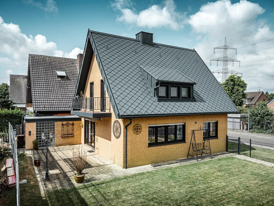 Ein Einfamilienhaus in Tönisvorst mit einem PREFA Dach aus Dachrauten 29×29 in P.10 Anthrazit, das ein herausstechendes Rautenmuster bietet. Schleppgauben, Kamin und der Eingangsbereich sind mit PREFALZ verkleidet, was zusammen mit Ablaufrohr und Hängerinne in P.10 Anthrazit dem Haus einen zeitlosen und eleganten Look verleiht. Das Backsteingebäude mit dem Aluminiumdach harmoniert mit der ruhigen, grünen Umgebung der Siedlung.