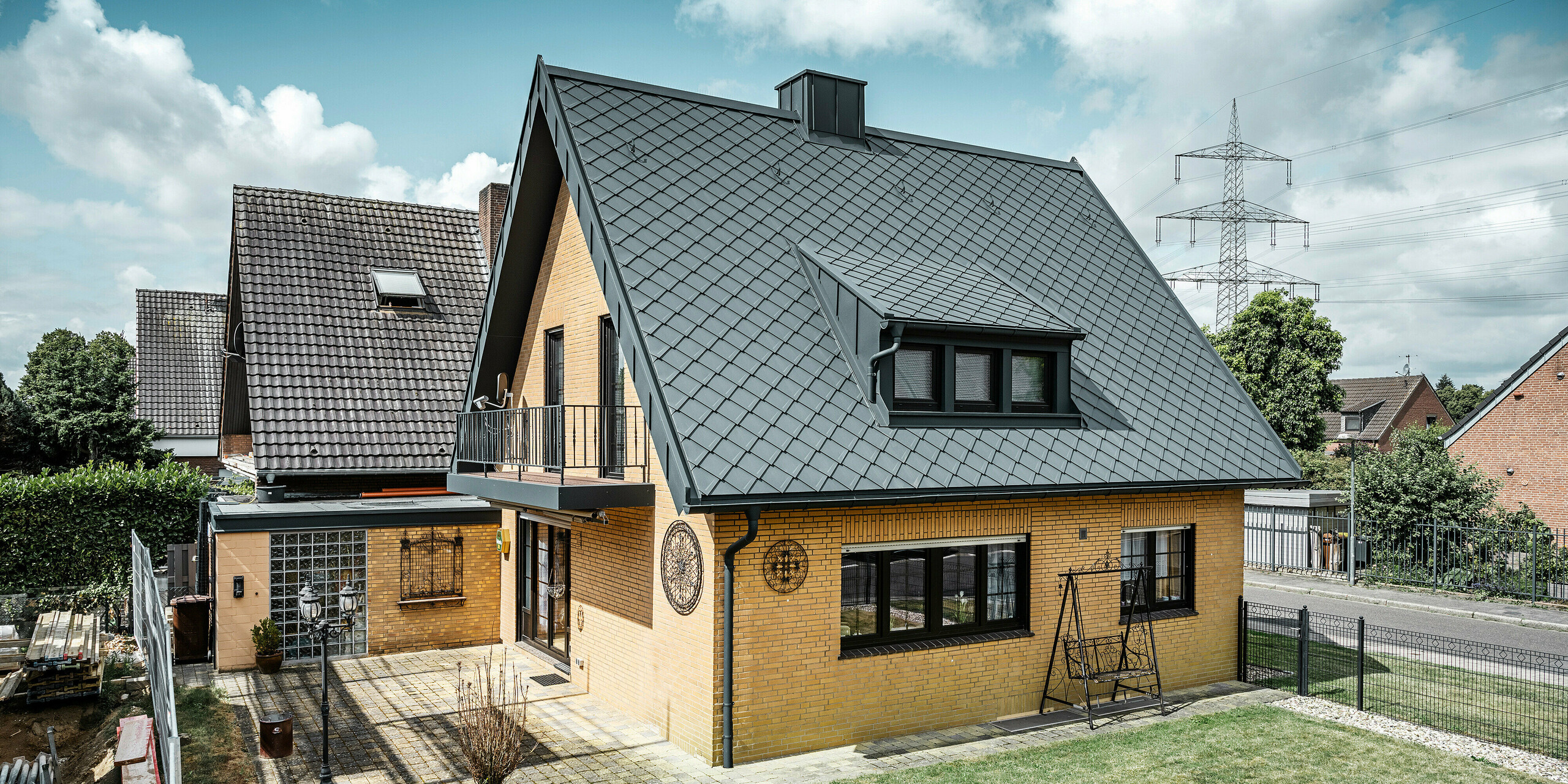 Ein Einfamilienhaus in Tönisvorst mit einem PREFA Dach aus Dachrauten 29×29 in P.10 Anthrazit, das ein herausstechendes Rautenmuster bietet. Schleppgauben, Kamin und der Eingangsbereich sind mit PREFALZ verkleidet, was zusammen mit Ablaufrohr und Hängerinne in P.10 Anthrazit dem Haus einen zeitlosen und eleganten Look verleiht. Das Backsteingebäude mit dem Aluminiumdach harmoniert mit der ruhigen, grünen Umgebung der Siedlung.