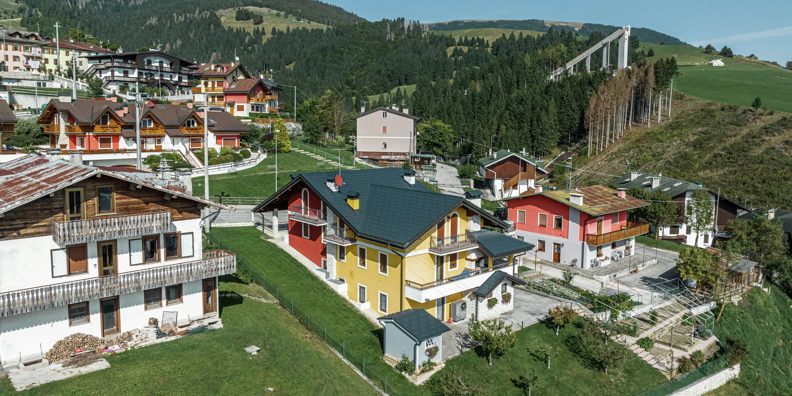 Maison individuelle idyllique à Gallio avec toiture moderne en aluminium PREFA composée de  R.16 P.10 en gris sombre. La couverture de toit en aluminium durable protège de manière fiable la maison de la neige, de la pluie et du vent - idéale pour les régions alpines. La façade jaune-rouge aux accents de pierre naturelle s'intègre harmonieusement dans l'environnement verdoyant. Des forêts denses et un tremplin de saut à ski en arrière-plan soulignent le caractère alpin typique de la région. Durable, robuste et esthétique - un toit parfait pour des générations.