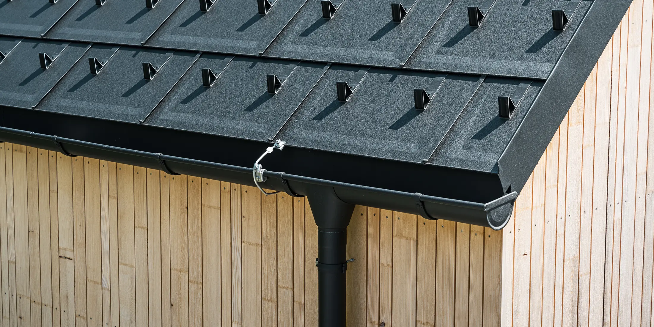 Nahaufnahme der PREFA Dachplatte R.16 mit Schneestoppern, Dachrinne und Ablaufrohr in Schwarz P.10 inkl. Blitzschutz und einer senkrechten Holzfassade. Die robusten PREFA Aluminiumprodukte schmücken das Satteldach eines Einfamilienhauses in Lustenau, Vorarlberg.