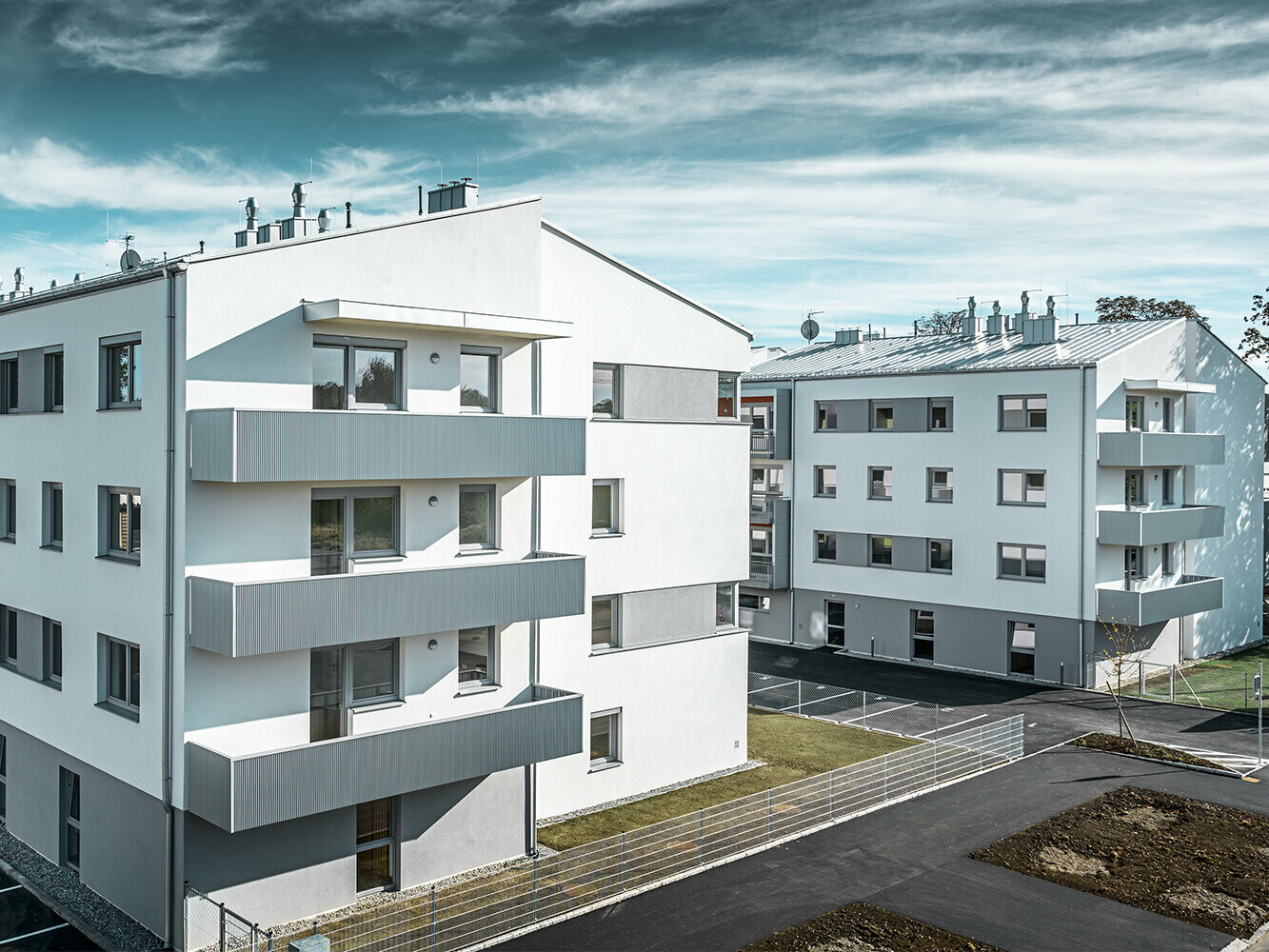 Perspektive aus der Ferne: Die moderne Wohnhausanlage der Glanzstoffsiedlung in St. Pölten präsentiert sich mit einer weißen Fassade und Balkonen, die mit dem PREFA Zackenprofil in Silbermetallic verkleidet sind. Die Aluminiumverkleidung der Balkone bildet einen harmonischen Blickfang in der Umgebung.