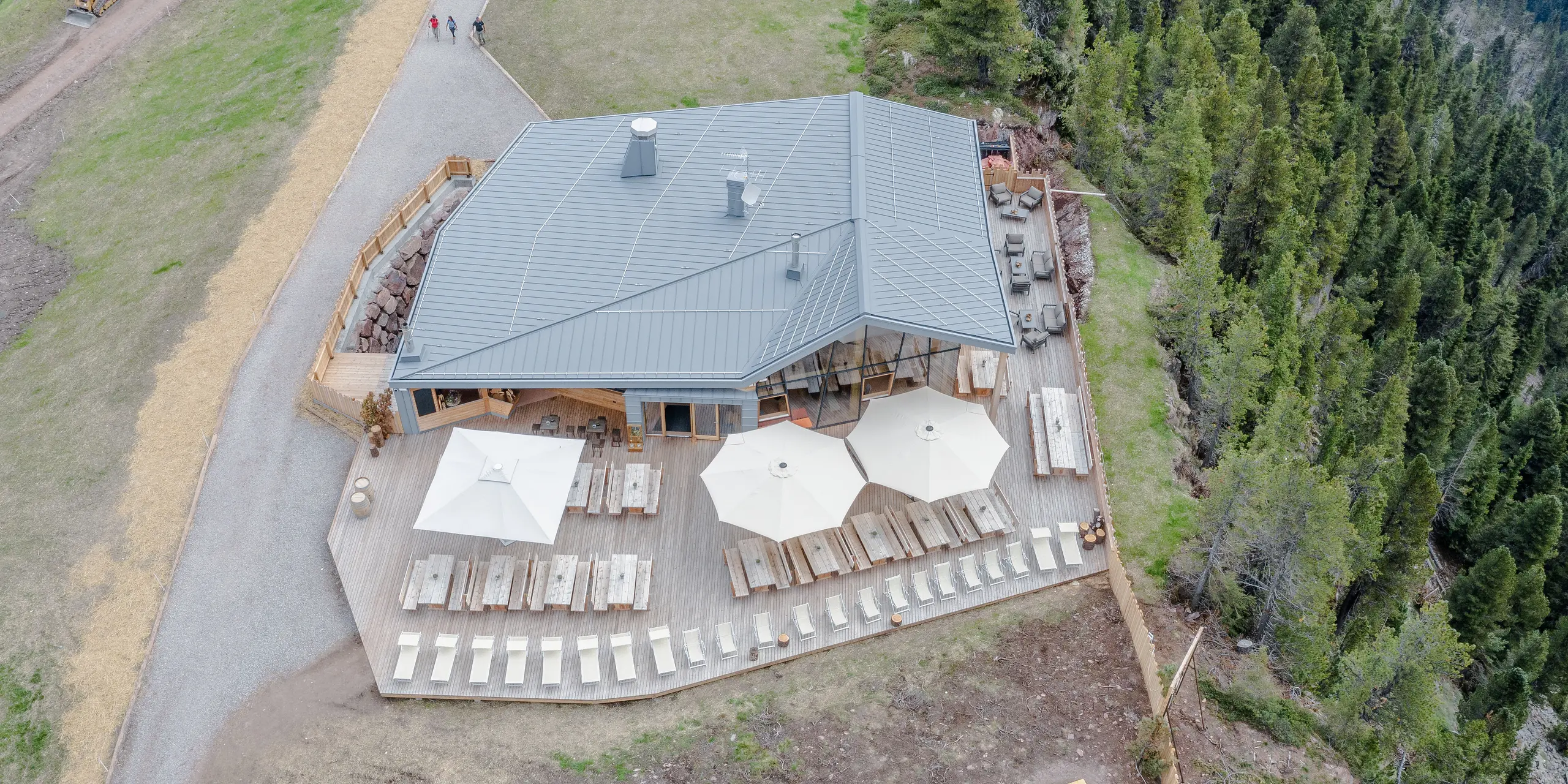 Luftaufnahme des Ristorante Busabelle, umgeben von sanften Wanderwegen und dichtem Wald. Die Architektur des Restaurants zeigt eine Kombination aus moderner hellgrauer PREFALZ Metallfassade und natürlichem Holz, die in die alpine Landschaft eingebettet ist. Die großflächige Terrasse ist ausgestattet mit Holzbänken und großen Sonnenschirmen, die einen komfortablen Aufenthaltsbereich für Gäste in dieser idyllischen Bergumgebung schaffen. Das Gebäude mit der Hülle aus Blech ist besonders robust und langlebig.