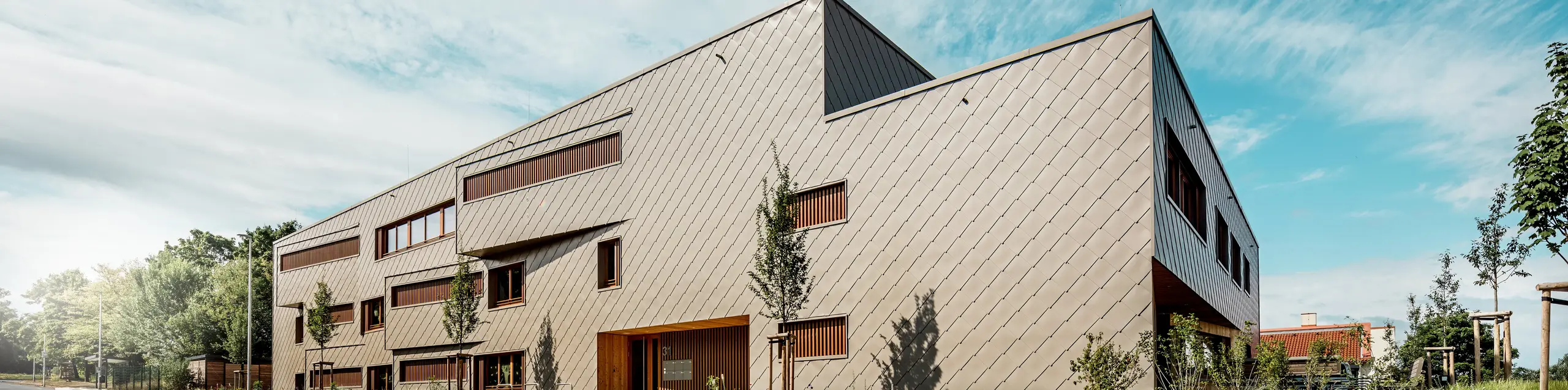 Vue en contre-plongée, la crèche d'Alzenau se distingue par ses lignes dynamiques et sa façade en aluminium PREFA bronze de haute qualité. La disposition diagonale des losanges de façade 44x44 confère au bâtiment une structure claire et une surface élégante et homogène. Des ouvertures de fenêtres décalées et des persiennes verticales en bois créent des accents rythmiques, tandis que le design minimaliste traduit une sobriété moderne. L'alliance de l'aluminium durable, du bois naturel et d'un concept d'aménagement paysager bien pensé le long de la rue souligne les exigences élevées en matière de durabilité et de qualité de construction. Cette image illustre avec brio comment une architecture fonctionnelle utilisant les produits PREFA peut être esthétiquement séduisante et définir de nouvelles normes en matière d'urbanisme.