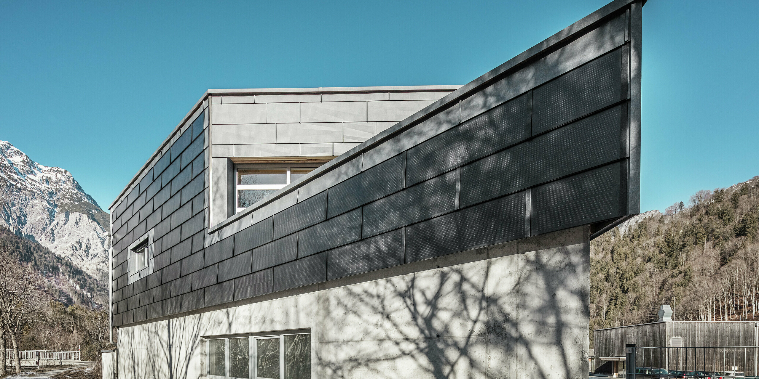 Le bâtiment de l'entreprise métallurgique RÜMA, dans le Vorarlberg, impressionne par sa façade moderne en tuiles solaires PREFA et béton apparent. Entouré de montagnes et d'arbres, dont les ombres se projettent sur le mur, il se dresse sous un ciel bleu et dégagé. L'angle pointu du bâtiment et les panneaux de façade PREFA FX.12 soulignent encore son allure imposante.