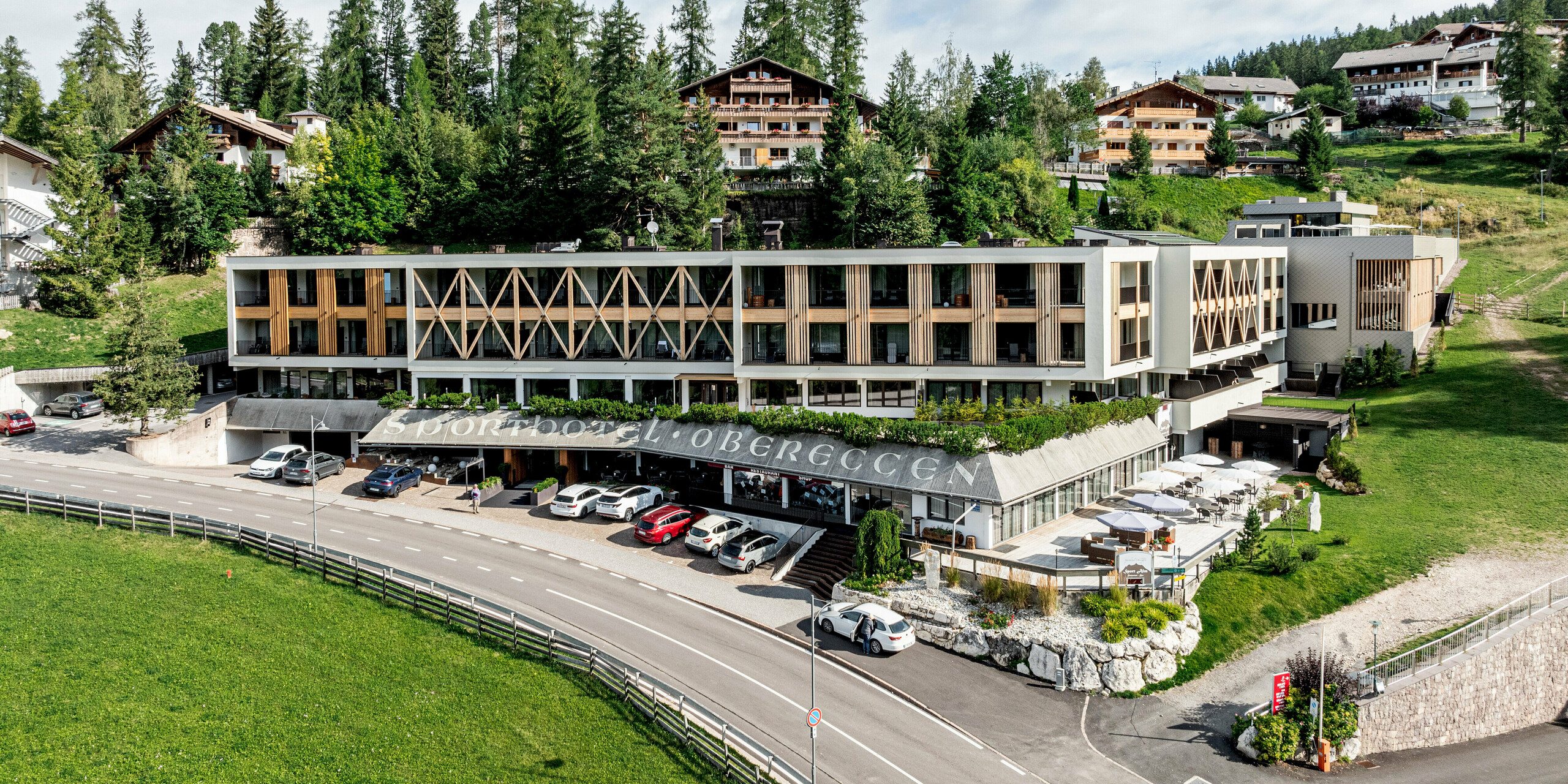 Gesamtansicht des Sporthotels Obereggen in Südtirol, harmonisch eingebettet in die alpine Landschaft mit Wiesen, Bäumen und umliegender Hangbebauung. Die moderne Hotelarchitektur wird durch die PREFA Aluminiumfassade mit Wandraute 29 × 29 in der Farbe Bronze geprägt, die dem Baukörper Struktur, Tiefe und Eleganz verleiht. Der warme Bronzeton bildet einen spannenden Kontrast zu Holz und Natur. Die langlebigen, witterungsbeständigen und wartungsarmen PREFA Aluminiumprodukte eignen sich ideal für hochwertige Architektur im alpinen Raum.