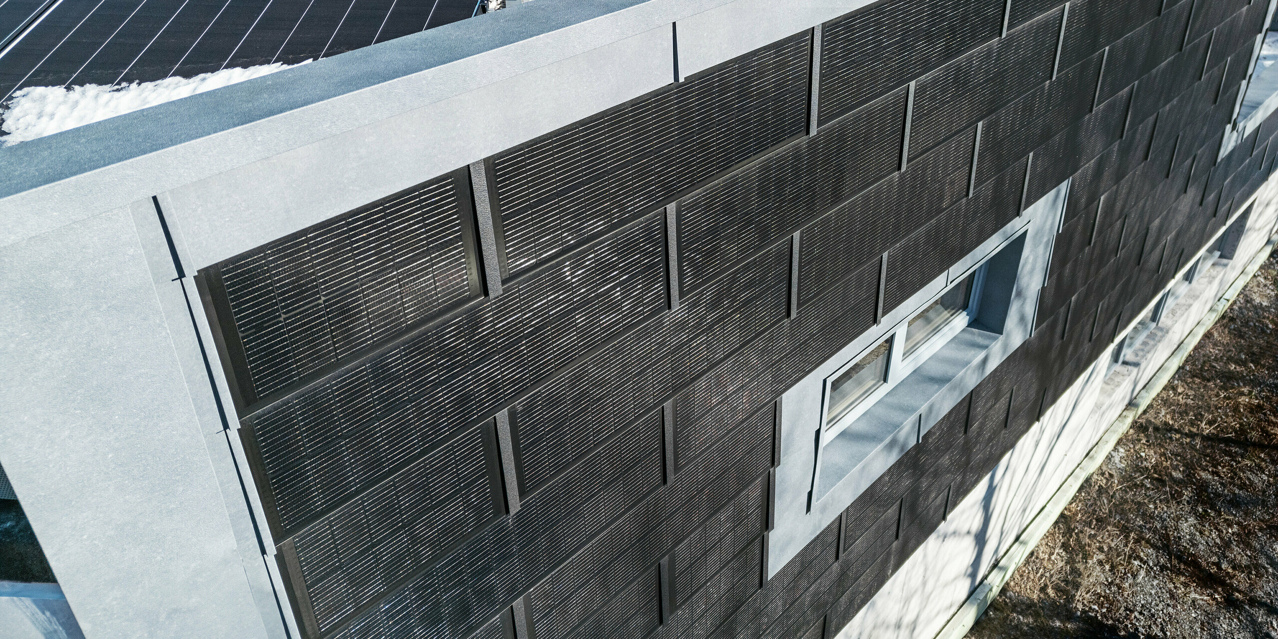 La vue de dessus montre la façade de l'entreprise métallurgique RÜMA, dans le Vorarlberg, avec ses panneaux de façade gris FX.12 et ses tuiles solaires PREFA innovantes avec modules photovoltaïques intégrés. Au premier plan, on aperçoit le toit plat partiellement enneigé, équipé de modules photovoltaïques supplémentaires.