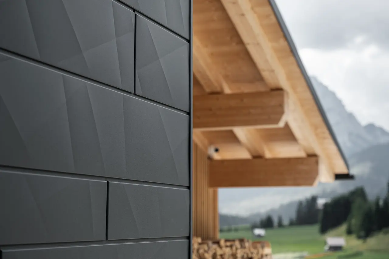 Nahaufnahme der Siding.X Fassadenpaneele in P.10 Schwarz von PREFA an der Casa Hoffe in Sappada, Italien. Das Fassadensystem aus Aluminium ist gekonnt in einem modernen, abstrakten Design angebracht, das einen ansprechenden Kontrast zu den sichtbaren Holzelementen des Dachstuhls bildet. Die innovative Fassadenlösung fügt sich nahtlos in das alpine Ambiente ein und steht für die Verbindung von Funktionalität, Beständigkeit und ästhetischer Baukultur.