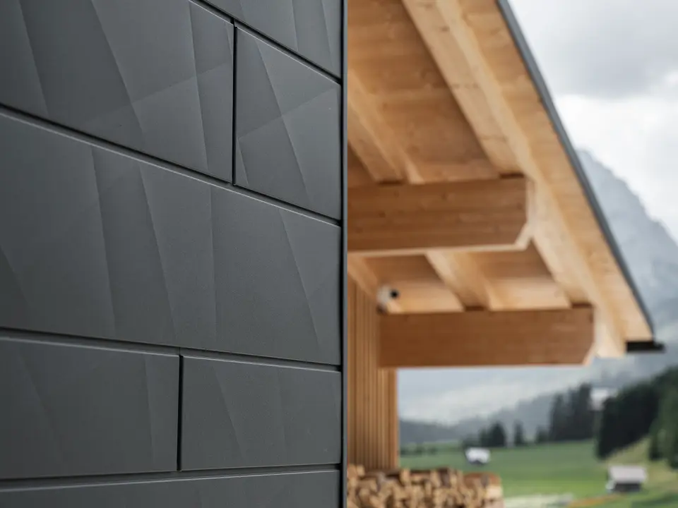 Detailfoto van de Siding.X gevelpanelen in P.10 zwart van PREFA aan Casa Hoffe in Sappada, Italië. Het gevelsysteem van aluminium is vakkundig aangebracht in een modern, abstract design, dat een aantrekkelijk contract vormt met de zichtbare houten elementen van de dakconstructie. De innovatieve geveloplossing past perfect in de alpiene omgeving en staat symbool voor de verbinding van functionaliteit, bestendigheid en esthetische bouwcultuur.