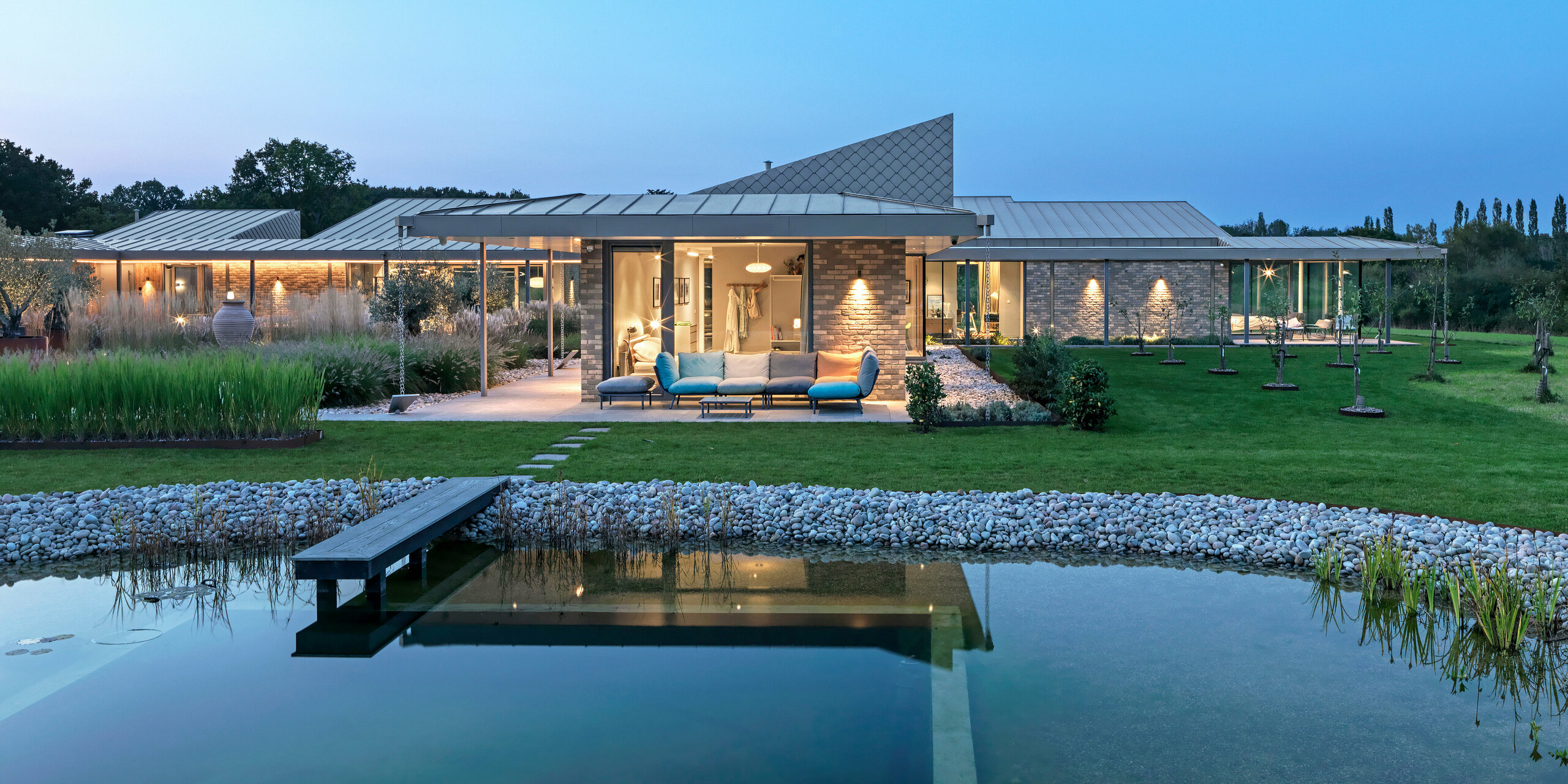 Vue nocturne d'une magnifique maison du West Sussex, avec toiture en aluminium PREFALZ et losanges de façade PREFA 20 × 20 P.10 bronze. Au premier plan, une piscine naturelle paysagée avec une passerelle en bois, derrière laquelle se trouve une spacieuse terrasse avec mobilier de jardin et une véranda éclairée. Les lignes épurées de l'architecture et la qualité des produits PREFA créent une élégante alliance de design, de fonctionnalité et de durabilité.