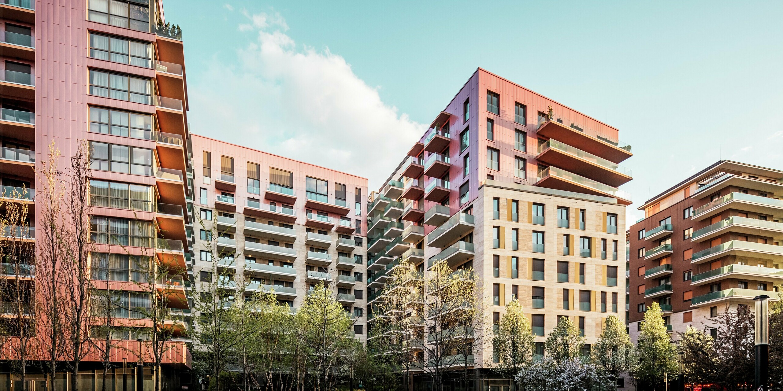 Vue d'ensemble du projet résidentiel Grand Corvin, dans le quartier revitalisé de Corvin à Budapest. Le revêtement de façade Siding PREFA, d'une couleur saumon métallisée, crée des accents visuels forts et met en valeur les étages supérieurs. Associé à la pierre naturelle et au verre, il crée un design architectural moderne et de haute qualité. Les panneaux en aluminium durables sont faciles d'entretien, résistants aux intempéries et s'intègrent harmonieusement à l'environnement urbain. La photo a été prise à l'heure dorée, ce qui permet à la lumière agréable et chaleureuse d'encadrer la scène.