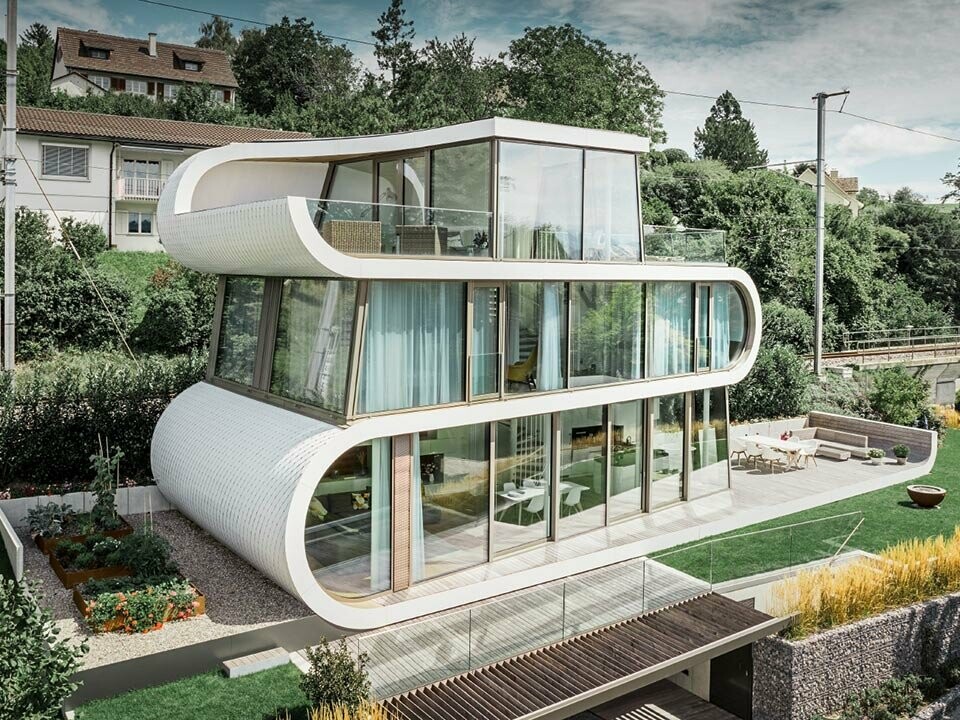 Opname van een modern flexhouse aan de Zürichsee; Dankzij de bijzondere vorm valt het flexhouse altijd op. De verdiepingen lopen als gebogen witte band door het gebouw heen, de ronde gevelelementen zijn bekleed met aluminium schindels van PREFA in zuiver wit.