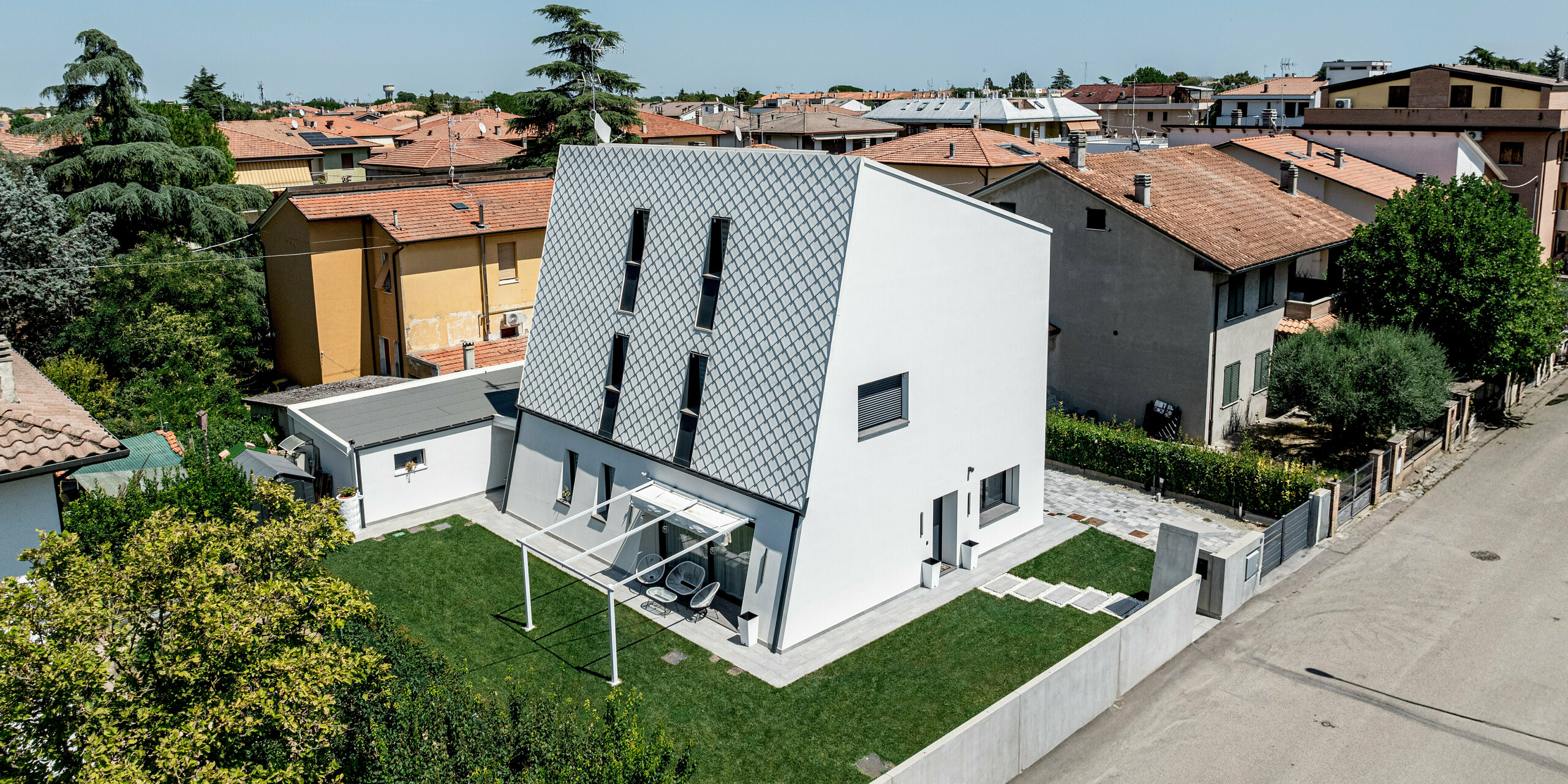 Schräg von oben aufgenommene Ansicht eines modernen Einfamilienhauses in Forlì, Italien, mit markanter, geneigter Hauptfassade aus PREFA Wandraute 29 × 29 in P.10 Reinweiß. Das präzise, diagonal verlaufende Rautenmuster aus Aluminium hebt sich deutlich von den umliegenden Ziegeldächern ab und wird zum architektonischen Blickfang. Klare Linien, schmale Fenster und der gepflegte Garten betonen den zeitgemäßen Charakter. Die PREFA Aluminiumfassade überzeugt durch Langlebigkeit, Witterungsbeständigkeit und eine dauerhaft hochwertige Optik.