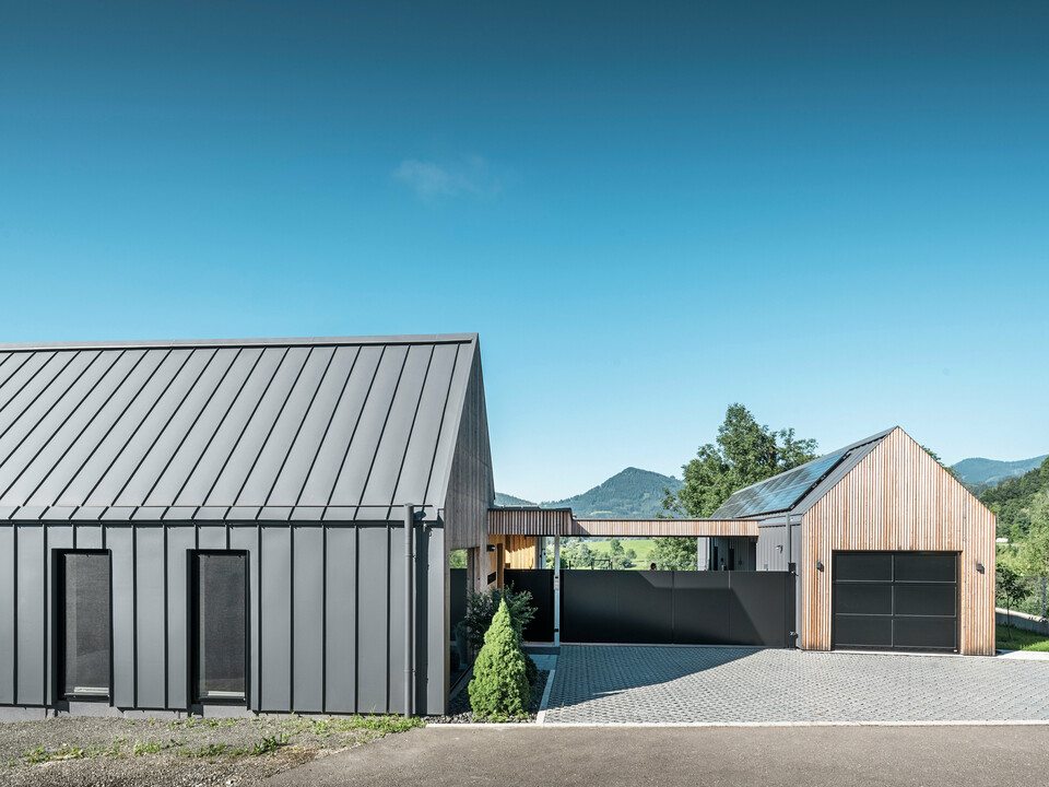 Vstupní areál rodinného domu s přilehlou garáží v rakouském Pogier uprostřed malebné krajiny. Střecha a fasáda obou budov jsou vybaveny odolným PREFALZ v P.10 tmavě šedé barvě, který vytváří moderní vzhled. Garáž se vyznačuje harmonicky integrovaným dřevěným obkladem, který hladce zapadá do okolní přírody. Dlážděné nádvoří a čisté linie designu doplňují estetiku nemovitosti, zatímco v pozadí zvlněné kopce dotvářejí obraz klidné zelené oázy v zeleném srdci Rakouska.