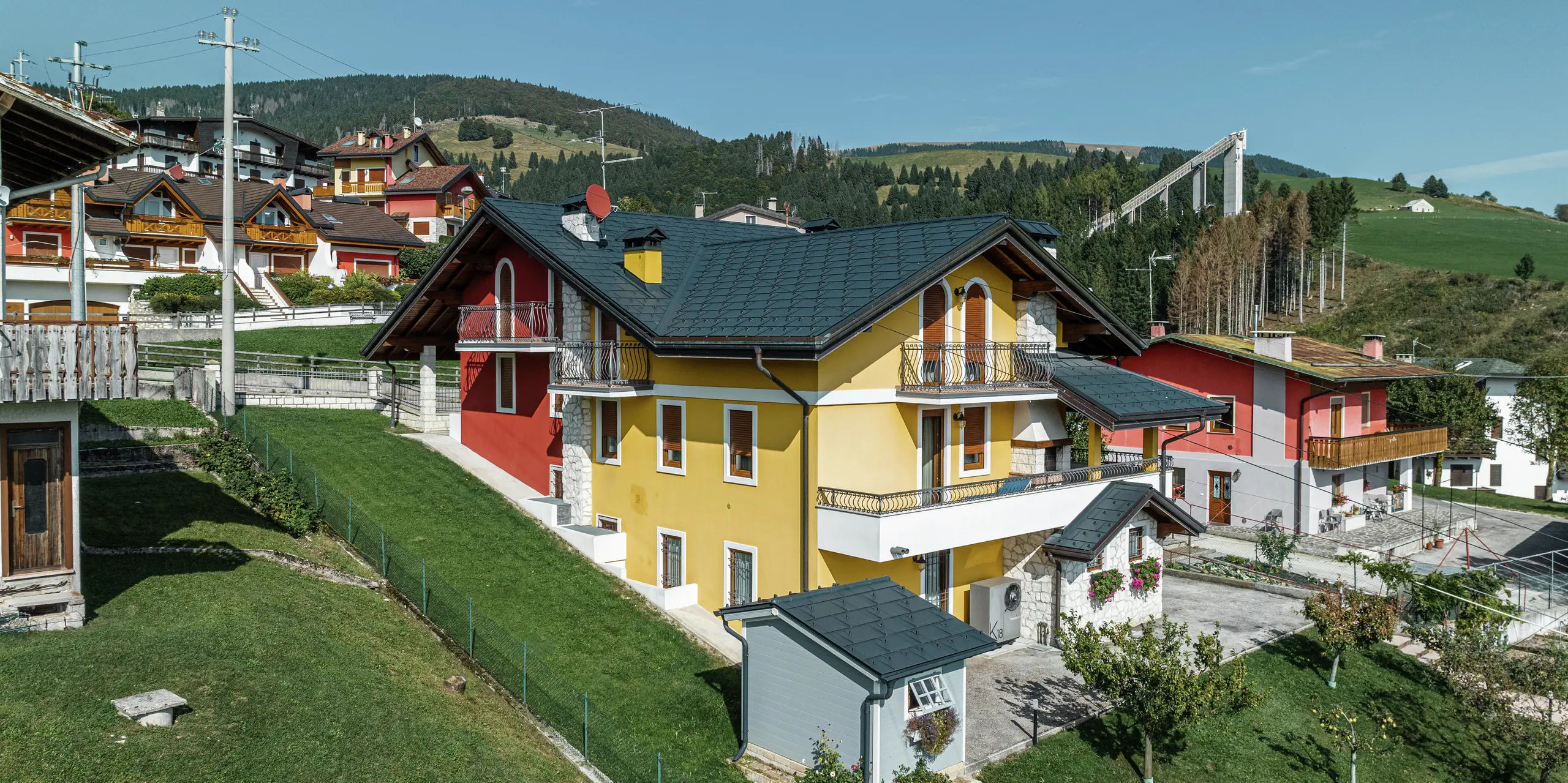 Maison individuelle à Gallio dans une architecture alpine traditionnelle avec une façade en plâtre jaune-rouge vif et des éléments en pierre naturelle. Le R.16 PREFA en P.10 gris sombre offre une résistance maximale aux intempéries et s'harmonise parfaitement avec l'environnement. Le système de gouttière avec gouttière suspendue et tuyau de descente PREFA – également en gris sombre P.10 – assure une protection fiable. Entourée de prairies verdoyantes et surplombant les montagnes, la maison allie esthétique, durabilité et fonctionnalité. En arrière-plan, vous pouvez voir le célèbre tremplin de saut à ski de la région.