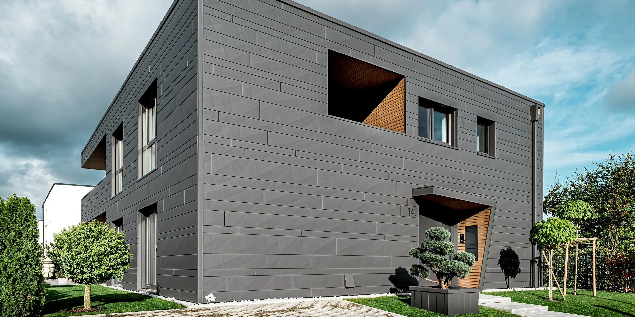 Cette maison individuelle moderne à Heidenheim présente une façade en aluminium entièrement revêtue de bardage Siding.X PREFA couleur P.10 gris sombre. Les arêtes vives et les largeurs variables des panneaux confèrent à la structure épurée du bâtiment une allure dynamique. L'entrée, habillée de bois, offre un contraste chaleureux avec l'élégance du métal. Un jardin paysager soigné occupe une place centrale. La robuste façade en aluminium PREFA et les tubes carrés intégrés garantissent une protection durable contre les intempéries, une grande stabilité et une esthétique résolument moderne.