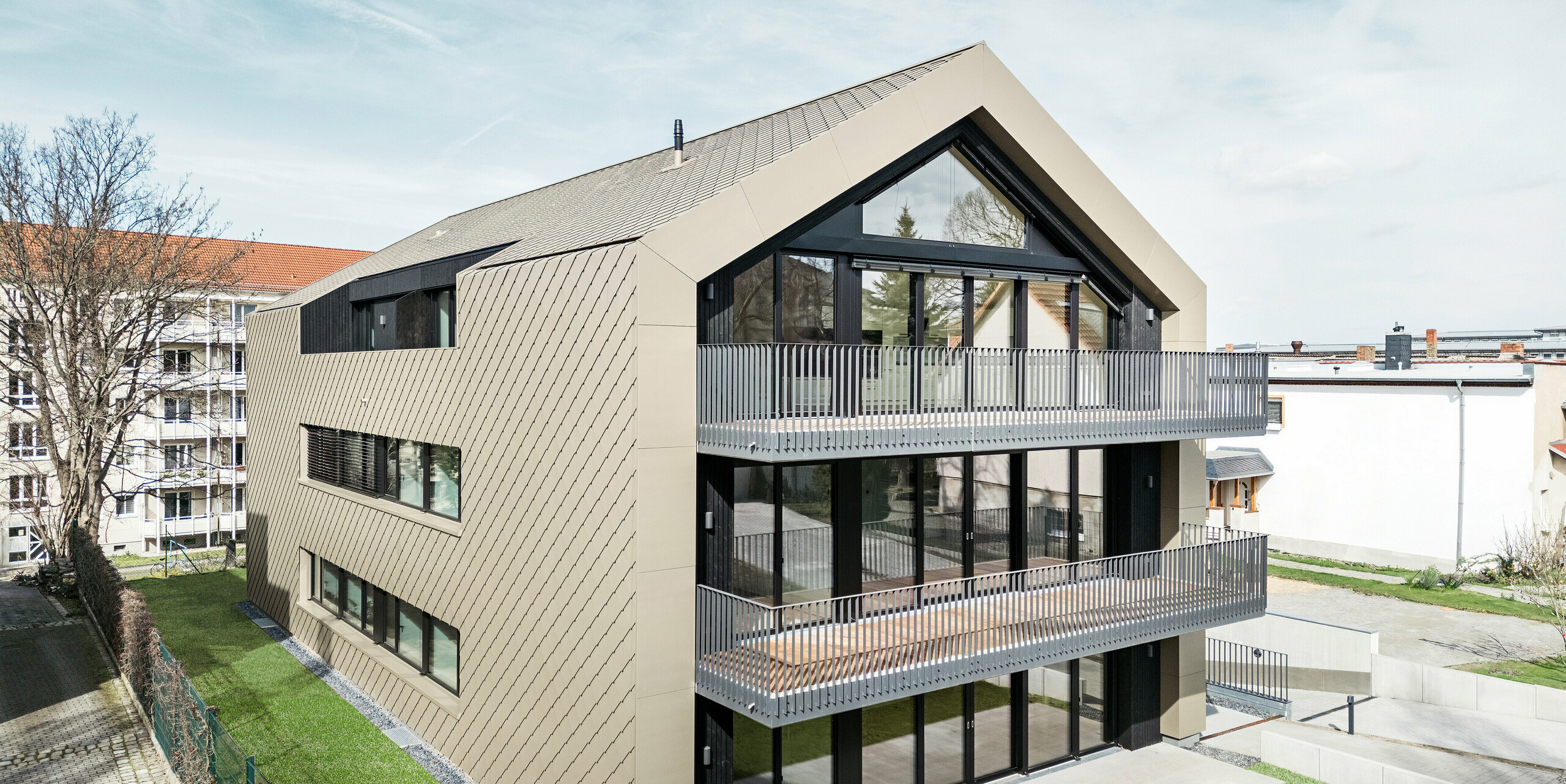Gartenansicht eines modernen Mehrfamilienhauses in der Chemnitzer Straße in Dresden. Der gepflegte Garten liegt vor einer eindrucksvollen Architektur. Die Fassade und das Dach des Gebäudes sind mit der langlebigen PREFA Aluminiumraute 29 × 29 in Bronze verkleidet, die dem Bauwerk eine elegante und zeitgemäße Optik verleiht. Das Bild zeigt die großzügigen Balkone, die großen Fensterfronten und die harmonische Einbettung des Gebäudes in die Umgebung.