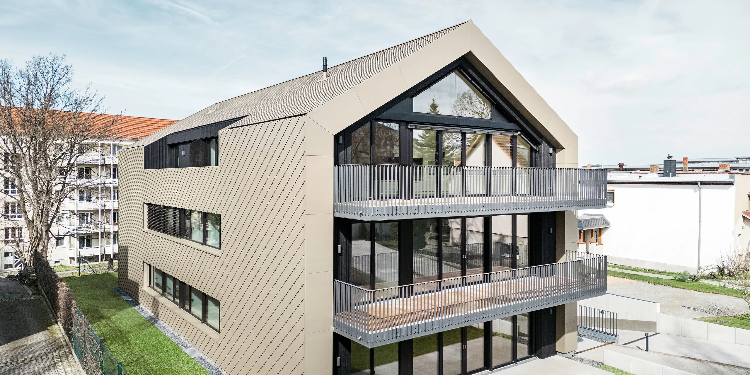 Vue sur le jardin d'un immeuble d'appartements moderne sur la Chemnitzer Straße à Dresde. Le jardin bien entretenu est entouré d'une architecture impressionnante. La façade et le toit du bâtiment sont revêtus du losange en aluminium durable PREFA 29 × 29 en bronze, qui confère à la structure un aspect élégant et contemporain. La photo montre les balcons spacieux, les grandes façades vitrées et l'intégration harmonieuse du bâtiment dans l'environnement.