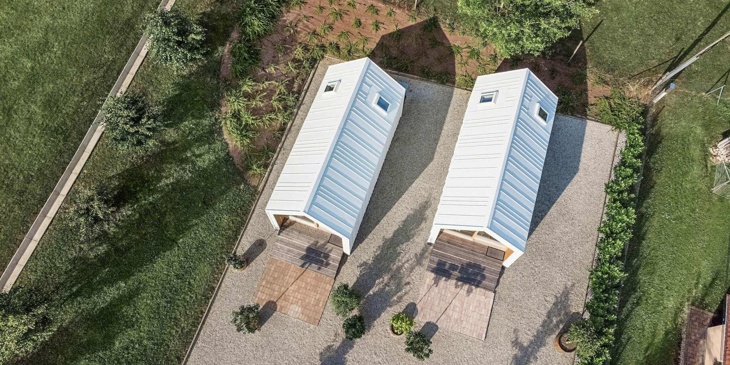 Vue aérienne des deux petites maisons du complexe de vacances « Pri Momi » à Kančevci, en Slovénie, conçues avec un revêtement de toit et de façade PREFALZ en blanc pur P.10. La photo montre la disposition symétrique des deux bâtiments avec leur coque blanche en aluminium robuste de haute qualité et les espaces verts environnants. Les lignes claires et l'architecture moderne des maisons sont soulignées par l'exécution précise du revêtement en aluminium. Les sentiers environnants et l'environnement naturel s'intègrent harmonieusement dans l'ensemble et invitent les visiteurs à s'attarder.