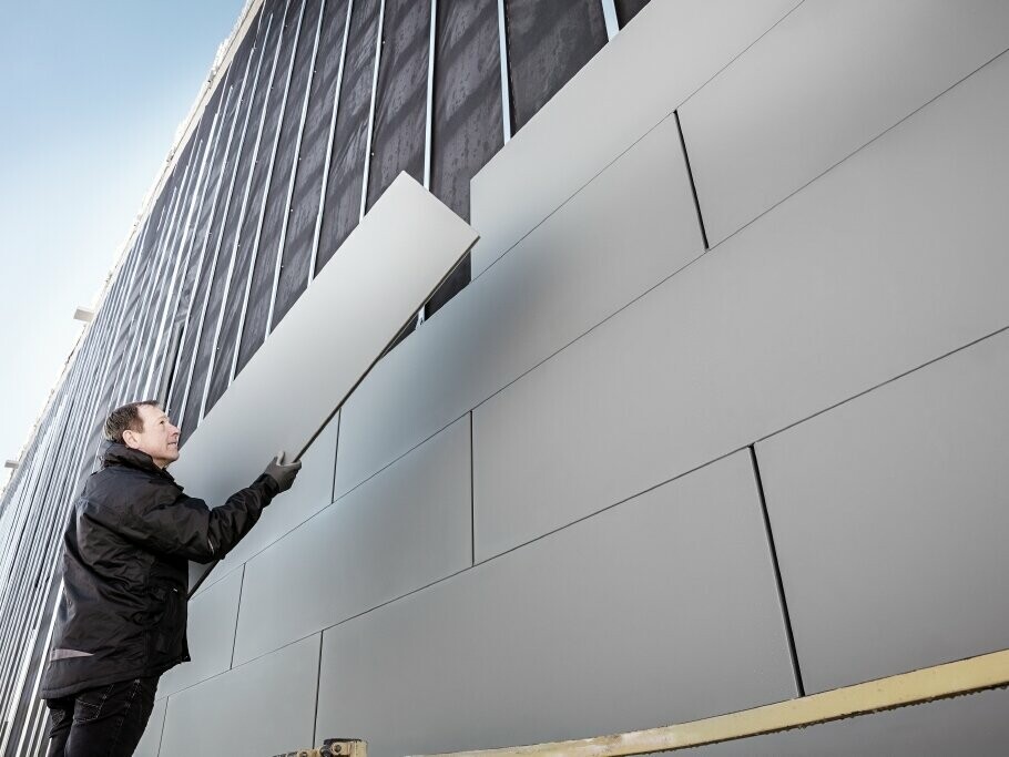 Fotos einer teilweise fertiggestellten Fassade, die mit PREFA Sidings in der Breite 500 verkleidet ist. Der Spengler verlegt ein weiteres Fassadenpaneel. Das Bild ist von unten aufgenommen. 