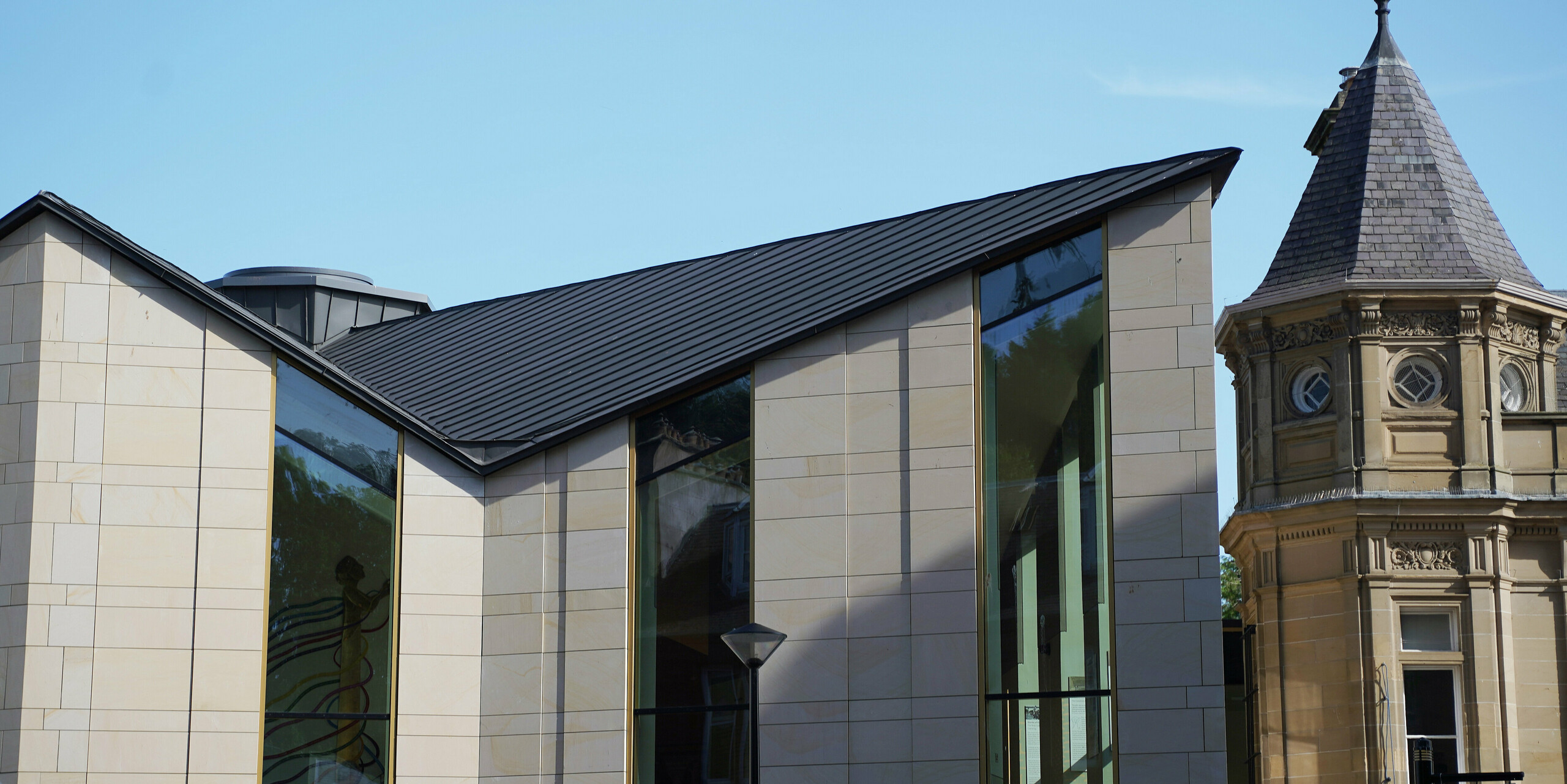 Seitenansicht des Museums 'The Great Tapestry of Scotland' in Galashiels, das sich durch ein modernes PREFALZ Dach in P.10 Zinkgrau auszeichnet. Das Dach mit seinen klaren, geometrischen Linien bildet einen reizvollen Kontrast zu den traditionellen Sandsteinfassaden der benachbarten Gebäude. Die einzigartige Dachkonstruktion mit den markanten Kanten verleiht dem Gebäude ein zeitgenössisches Flair und unterstreicht die Verbindung von historischer Architektur und modernem Design.