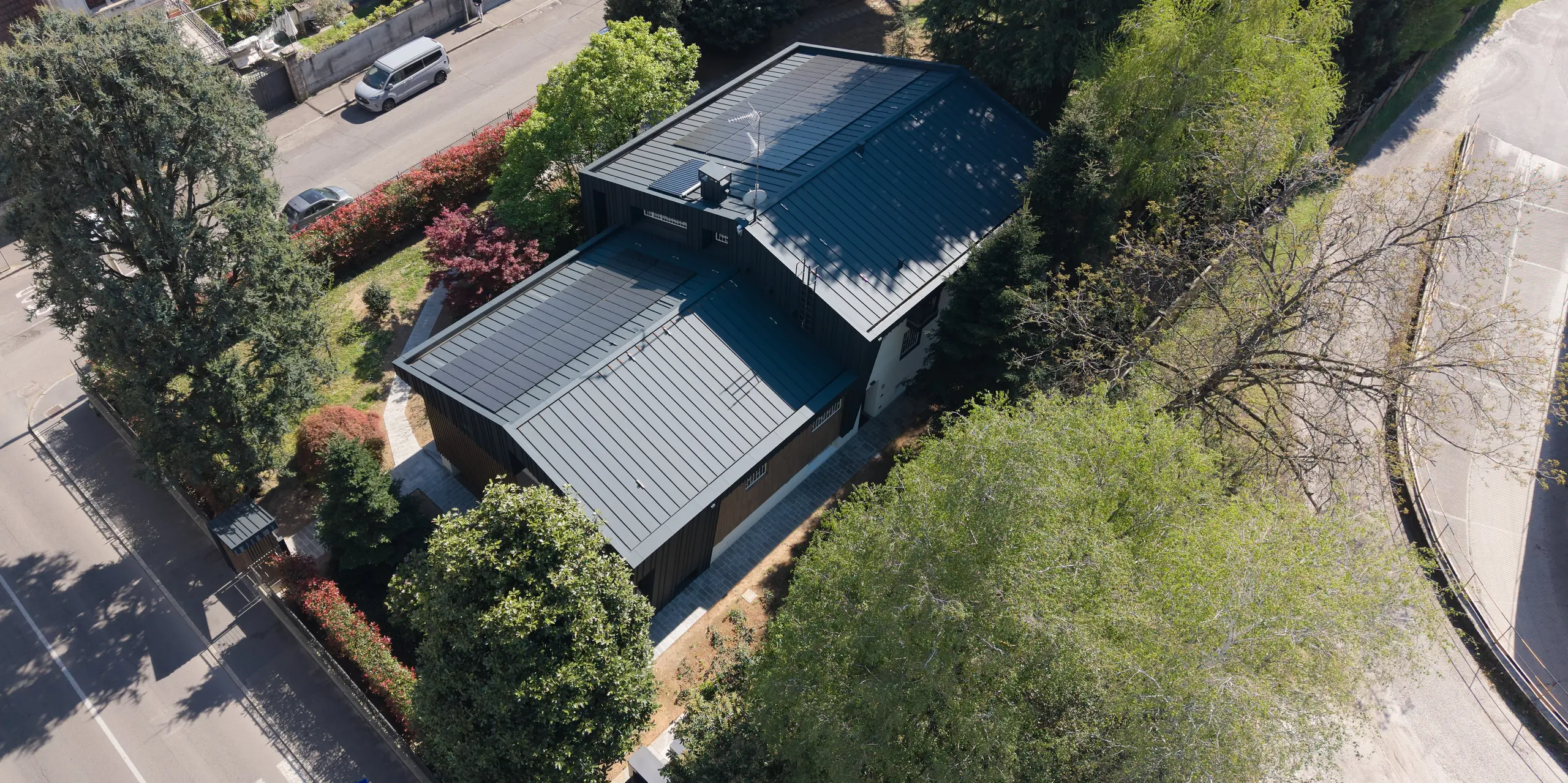 Die Luftaufnahme zeigt die moderne Villa Castellanza mit ihrer markanten Gebäudehülle aus PREFA Aluminium. Besonders auffällig sind das elegant gefalzte PREFALZ Dach und die nahtlos integrierte PREFALZ Fassade in P.10 Anthrazit. Sie verleihen dem Wohnhaus eine klare, zeitgemäße Architektur. Die großflächig eingebundenen PREFALZ Solarmodule fügen sich harmonisch in die Dachlandschaft der beiden Satteldächer ein und unterstreichen das nachhaltige Energiekonzept. Umgeben von altem Baumbestand entsteht ein spannender Kontrast zwischen Natur und moderner Architektur. Die langlebigen und wartungsarmen PREFA Aluminiumlösungen verbinden anspruchsvolles Design, Funktionalität und Energieeffizienz auf höchstem Niveau.