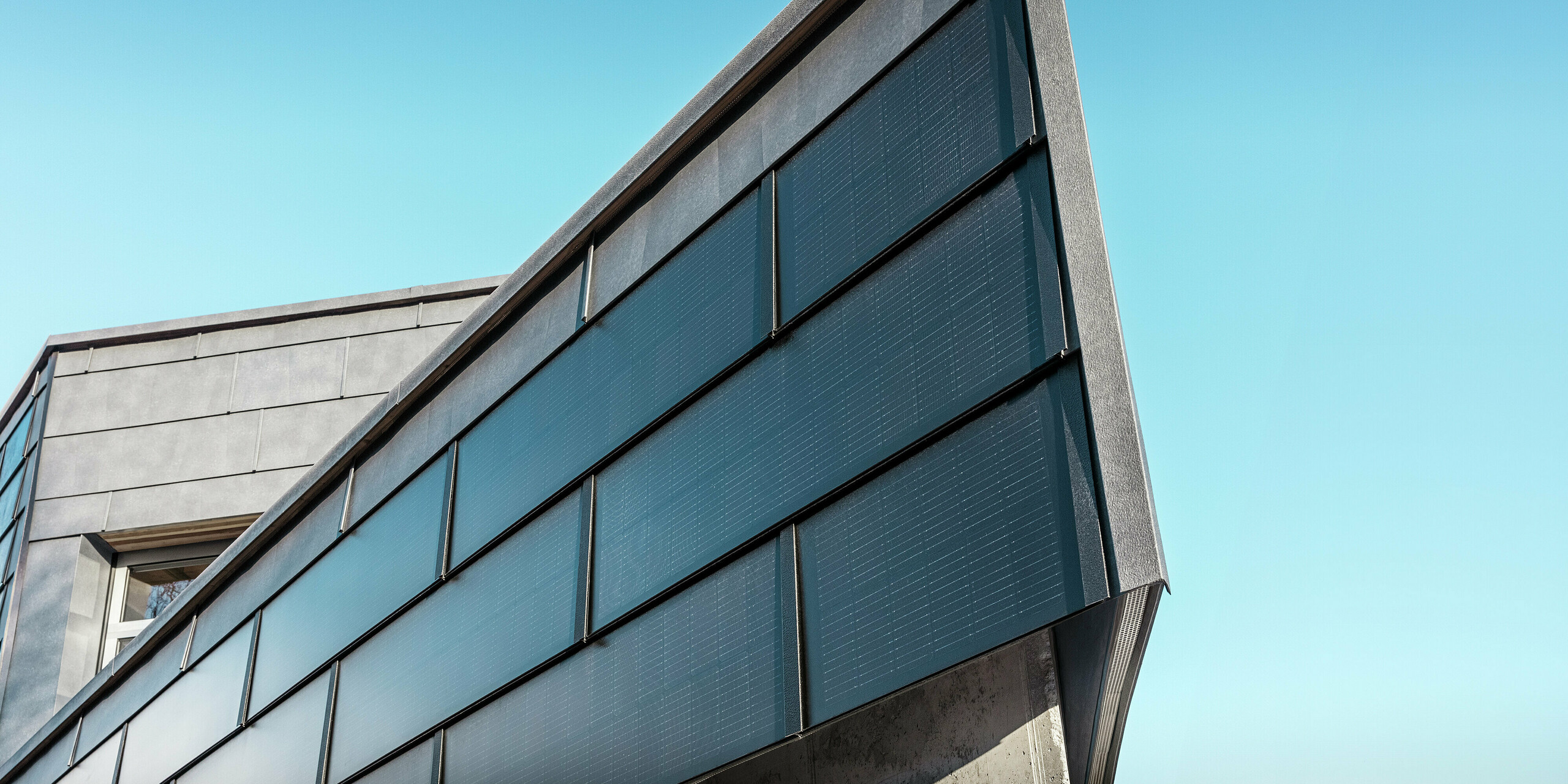 La façade moderne de l'entreprise de couverture RÜMA, dans le Vorarlberg, est dotée de tuiles solaires PREFA en aluminium et de modules photovoltaïques intégrés, associés à des panneaux de façade PREFA FX.12 pour une production d'énergie durable et un design de façade dynamique. La photo montre un angle du bâtiment vu de près. Le ciel bleu éclatant est visible en arrière-plan.