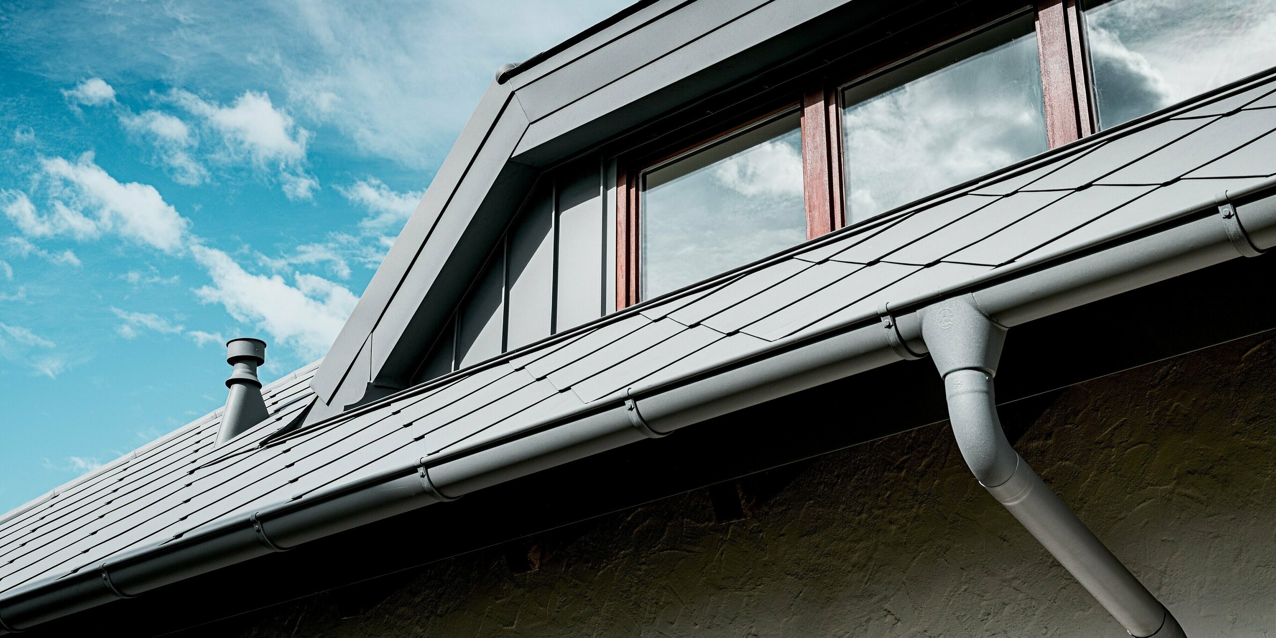 Das Bild zeigt eine detaillierte Ansicht der Trapezgaube eines Einfamilienhauses, eingedeckt mit PREFA Dachrauten 44 × 44 in P.10 Hellgrau. Die klaren Linien der Gaubenverkleidung gehen harmonisch in die strukturierte Rautenfläche über und verleihen dem Dach eine moderne, hochwertige Optik. Im Vordergrund ist die PREFA Dachentwässerung mit Hängerinne und Ablaufrohr sichtbar, sauber integriert in das langlebige Aluminiumdach. Vor dem blauen Himmel wirkt das gesamte Dachsystem besonders robust, präzise verarbeitet und widerstandsfähig.