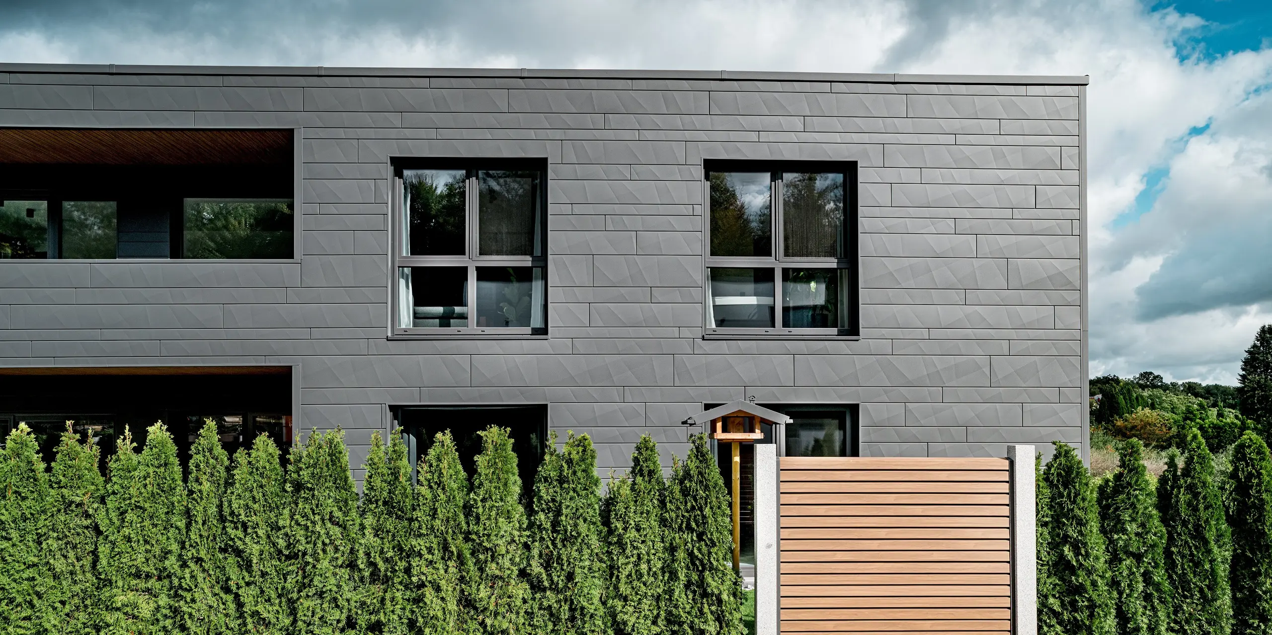 Vue latérale d'une maison individuelle moderne à Heidenheim, revêtue d'une façade entièrement habillée de panneaux Siding.X PREFA, coloris P.10 gris sombre. Les panneaux d'aluminium horizontaux, de largeurs variables et aux bords marqués, créent une surface texturée et dynamique. Au premier plan, une haie dense de thuyas à feuillage persistant s'étend sur le paysage, tandis qu'une clôture en bois apporte une touche chaleureuse à l'architecture épurée de la façade. De larges fenêtres reflètent l'environnement et soulignent la pureté des lignes. La surface en aluminium PREFA, résistante et durable, garantit une protection optimale et une élégance contemporaine.