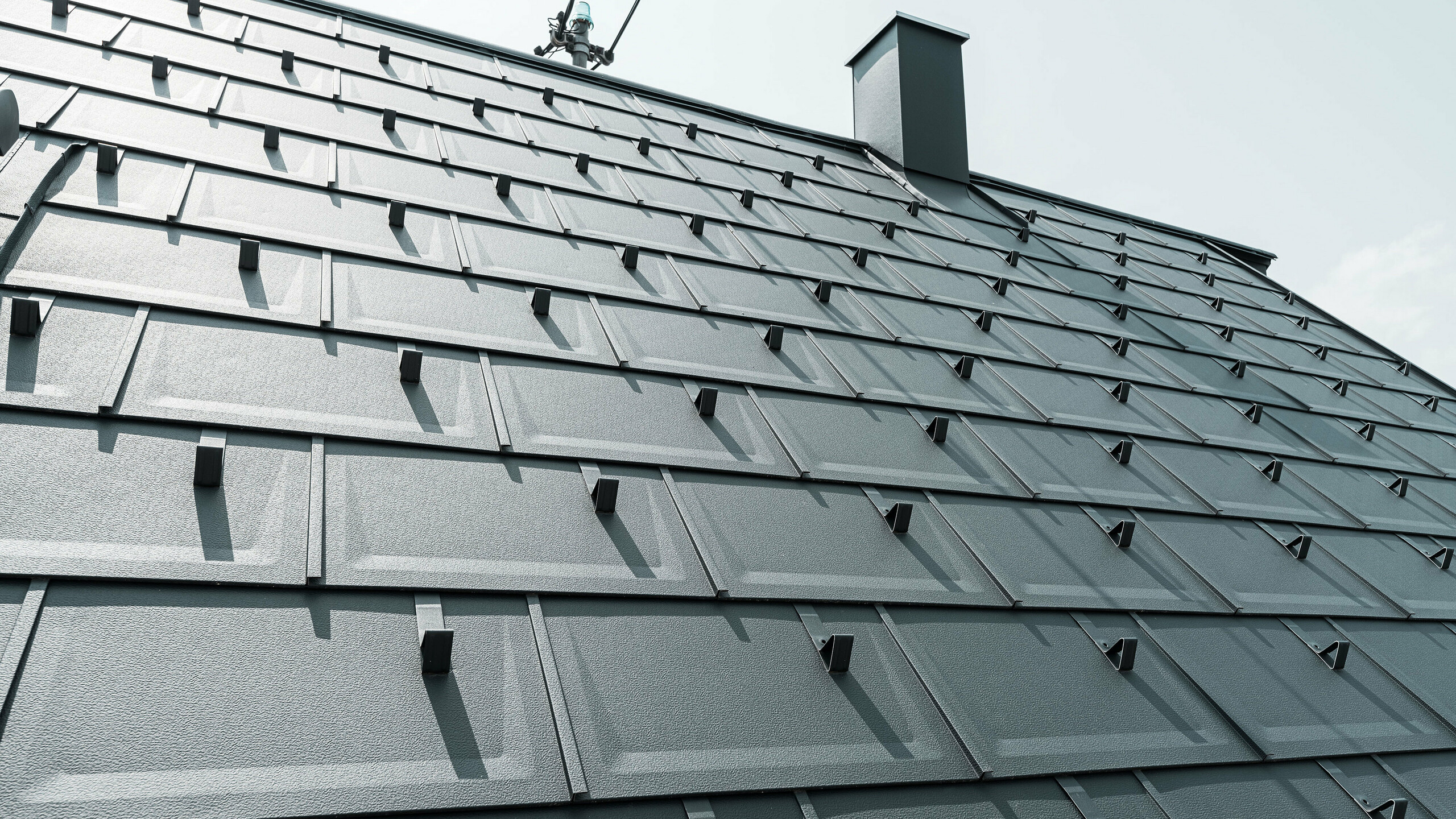 Detailaufnahme der PREFA Dachplatte R.16 in der Farbe P.10 Anthrazit. Am Dach sind PREFA Schneestopper und ein Rauchfang zu sehen.