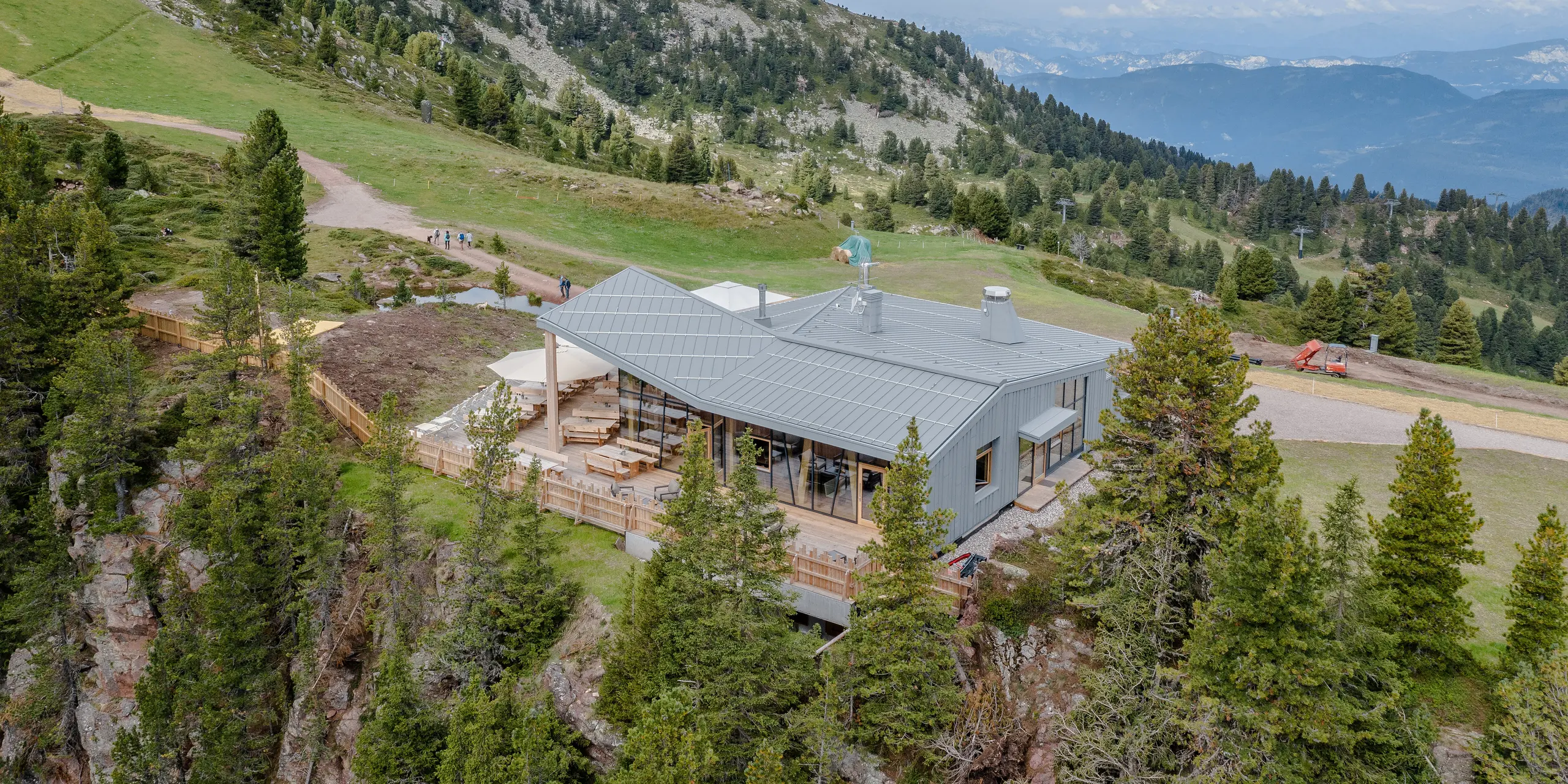 Eindrucksvolle Aufnahme des Ristorante Busabelle, gelegen in einer hügeligen und waldreichen Landschaft mit weitreichendem Blick auf entfernte Bergketten. Das Restaurant, mit seiner charakteristischen hellgrauen PREFALZ Dach- und Fassadenverkleidung, bietet eine breite Terrasse und ist umgeben von natürlichen Wegen, die sich ideal für Spaziergänge in der malerischen Umgebung eignen. Dank der Aufnahme durch eine Drohne ist die wunderschöne Berglandschaft im Hintergrund einwandfrei zu erkennen.