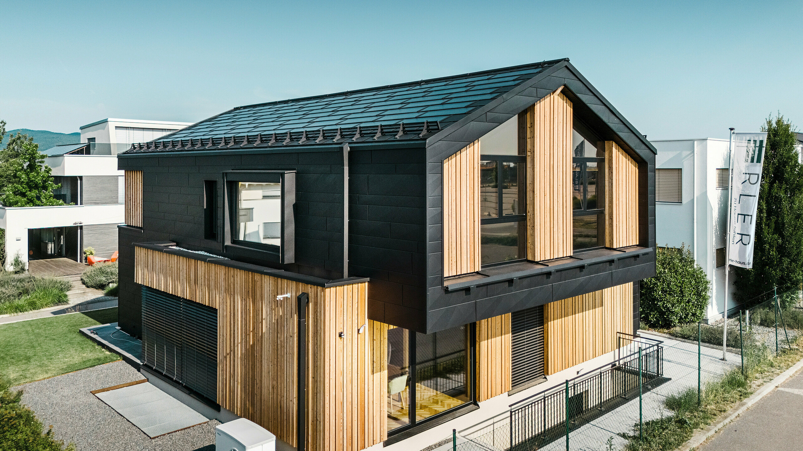 Façade de la maison modèle Autark dans le centre de maisons préfabriquées Blaue Lagune, Vösendorf (AT). Le toit est recouvert de tuiles solaires PREFA noirs et de R.16 avec un revêtement P.10 de haute qualité. La façade est constituée d'une combinaison d'éléments verticaux en bois et de PREFA Siding.X en P.10 noir, qui confère au bâtiment un caractère moderne et durable. Le drapeau de la société Erler Bau GmbH flotte sur le bord droit de l'image.
