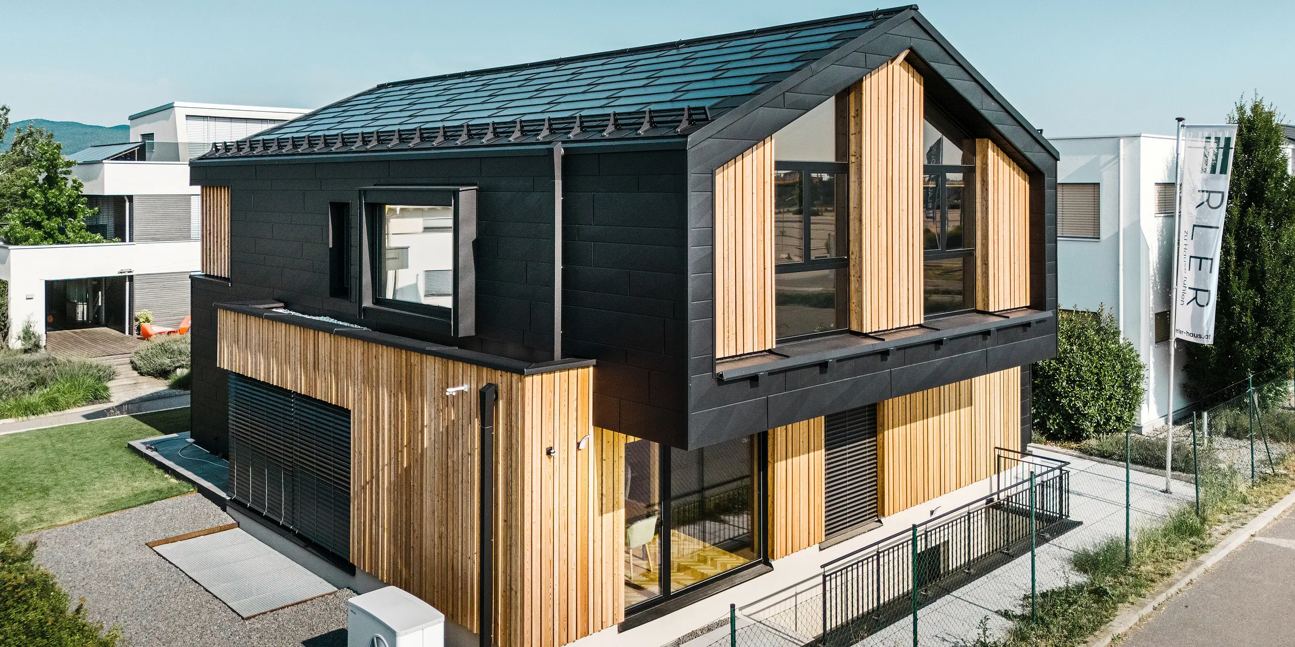 Voorzijde van het modelhuis Autark in het prefabhuiscentrum Blaue Lagune, Vösendorf (AT). Het dak is bedekt met zwarte PREFA solar-dakpan en dakpan R.16 met een hoogwaardige P.10-coating. De gevel bestaat uit een combinatie van verticale houten elementen en PREFA Siding.X in P.10 zwart, wat het gebouw een modern en duurzaam karakter geeft. Aan de rechterkant van de foto wappert de vlag van Erler Bau GmbH.