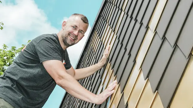 PREFA Handwerker mit grauem T-Shirt an einer PREFA Rauten-Fassade in Braun und Mayagold.