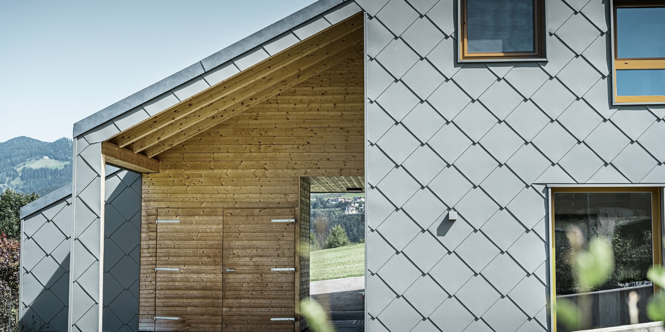 De overdekte toegangszone is voorzien van een horizontale houten bekleding; de rest van de gevel is bekleed met de grote PREFA gevellosange uit aluminium in lichtgrijs.